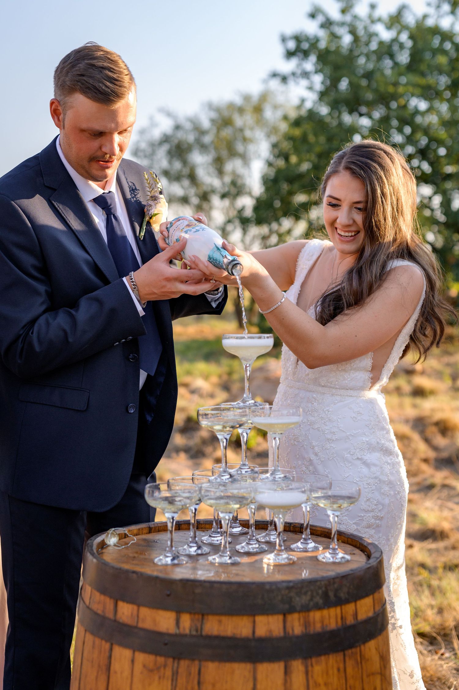 Couple pours champagne over stacked glasses on rustic wooden barrel outdoors.