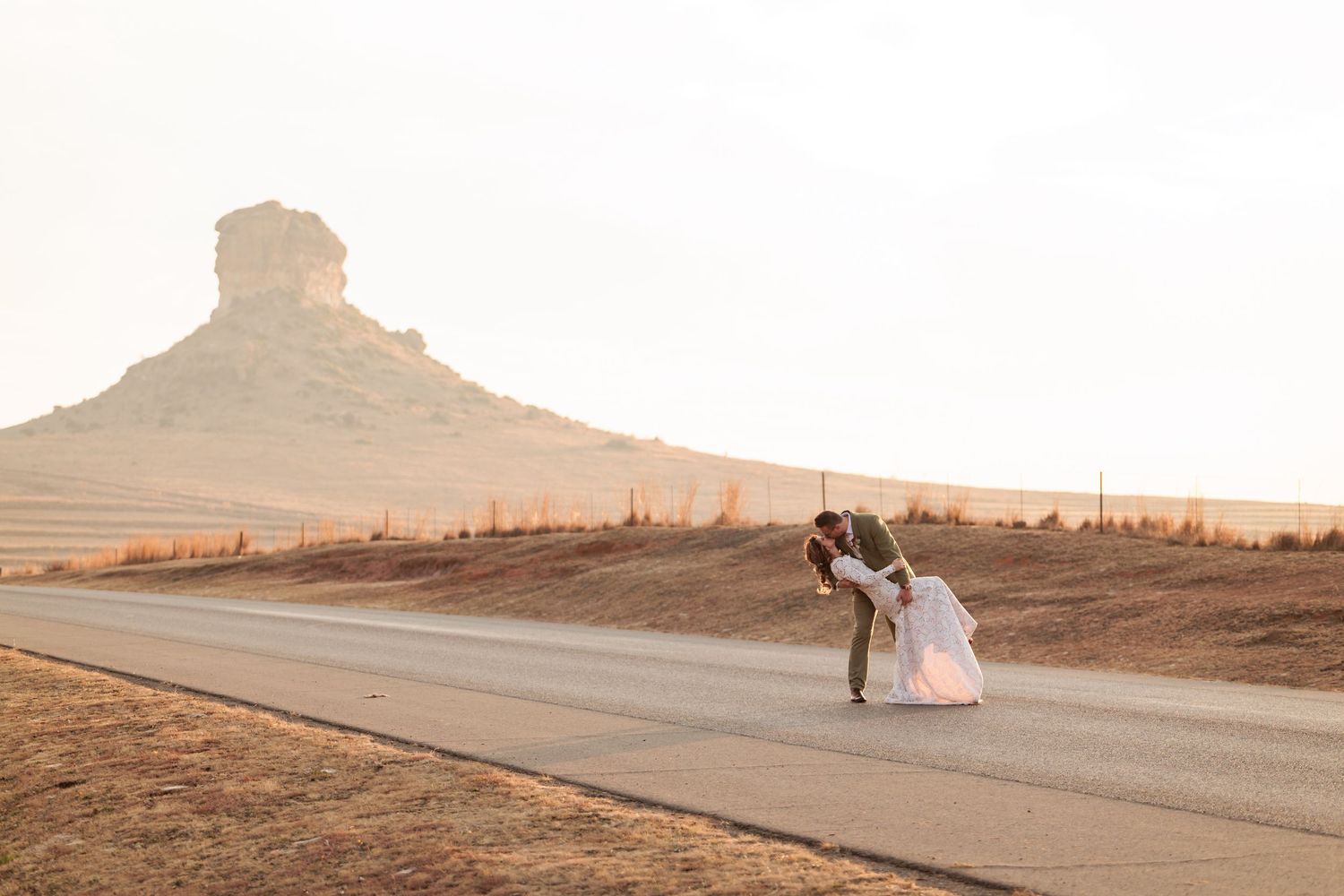 Couple sharing a romantic dip kiss on a desert road with a sandstone butte at golden hour.
