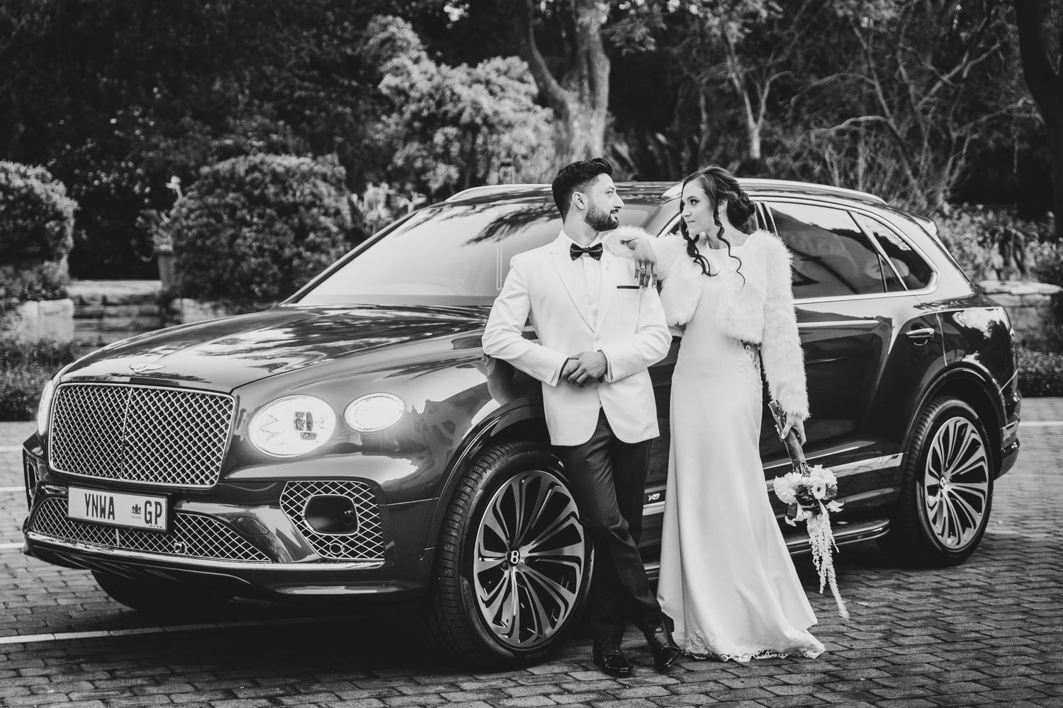Bride and groom posing in front of a luxury Bentley SUV on their wedding day in a scenic outdoor setting.
