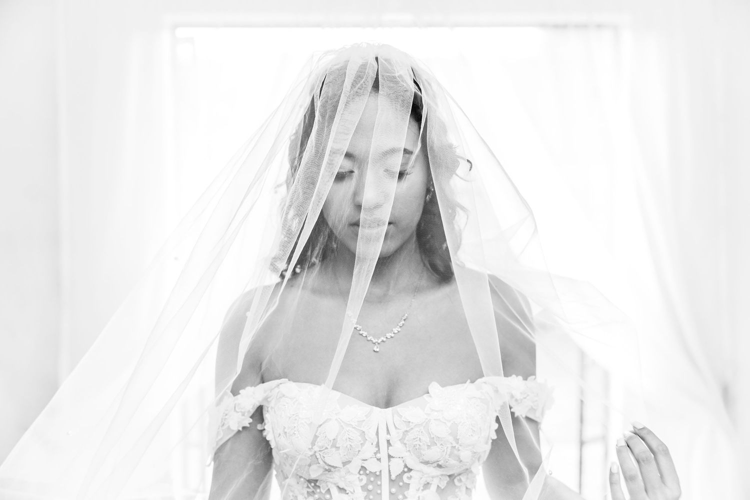 Black and white photo of a bride in lace gown and veil, looking down elegantly near a bright window.