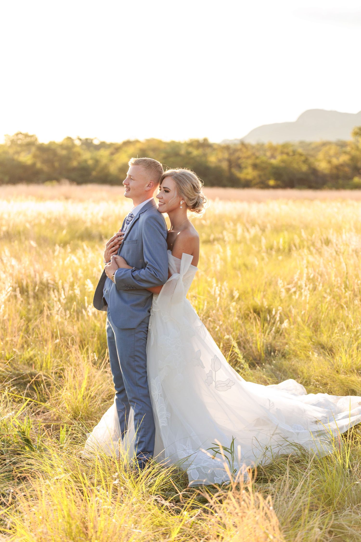 Newlywed couple embraces in golden sunlit field during sunset while bride's white dress flows in the breeze.