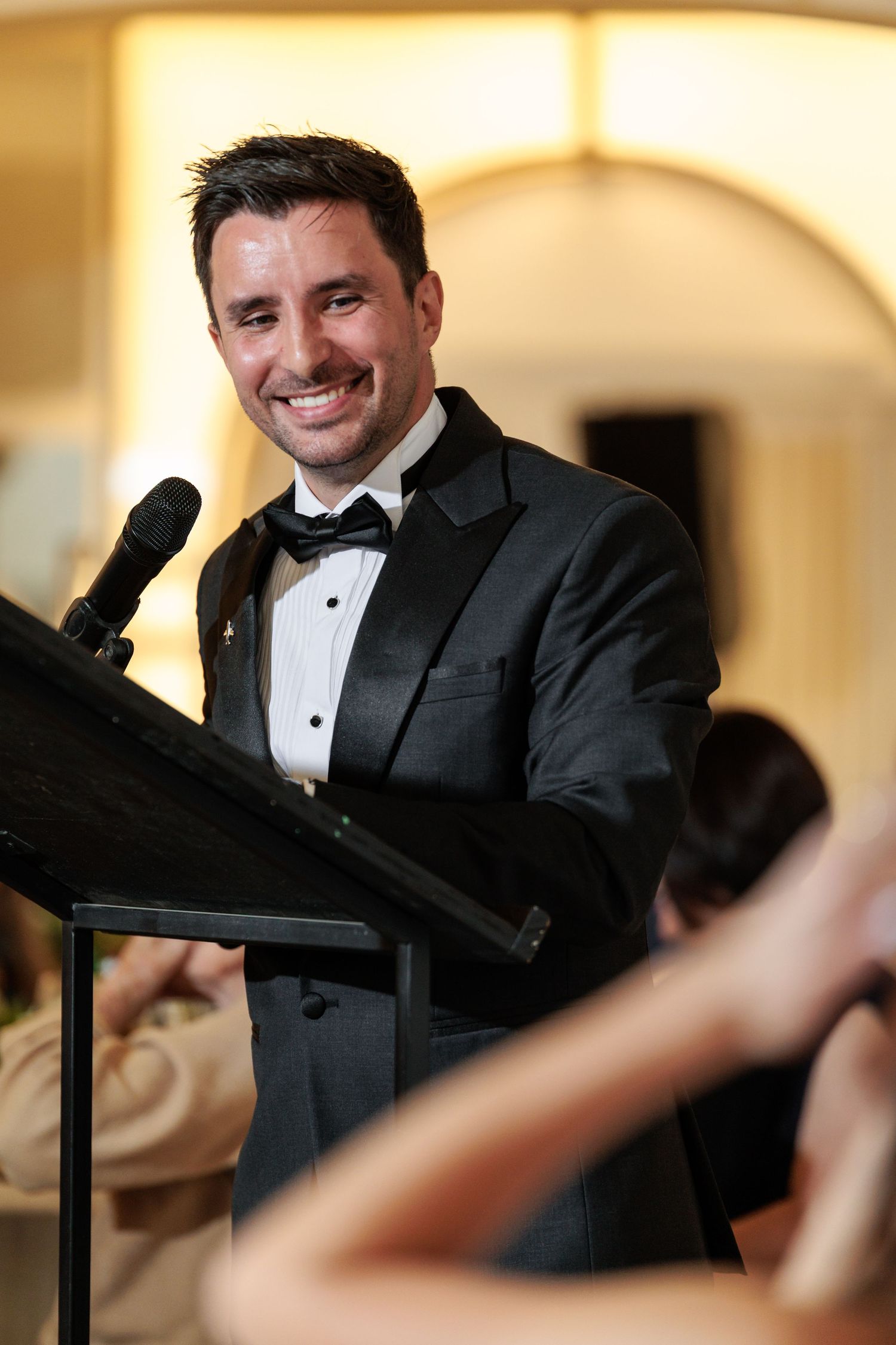 A person in a black tuxedo and bow tie leans on a railing in an elegant venue with arched architecture.