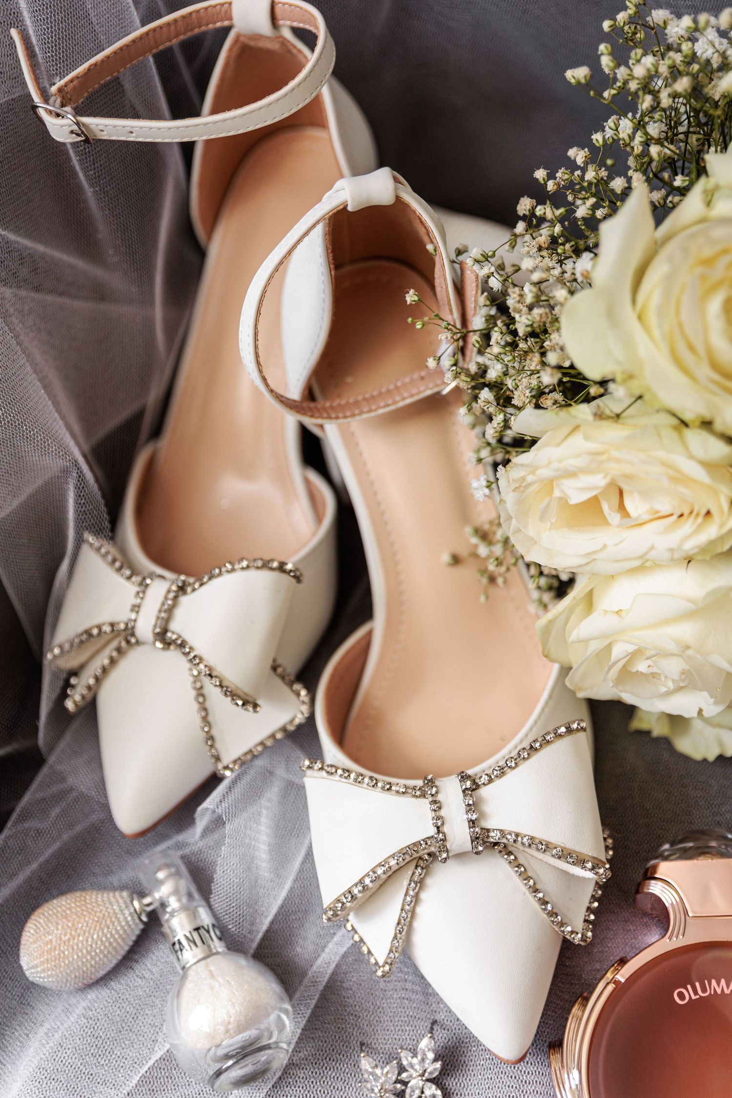 Elegant white bridal shoes with crystal bow embellishments displayed alongside delicate white roses and wedding accessories.