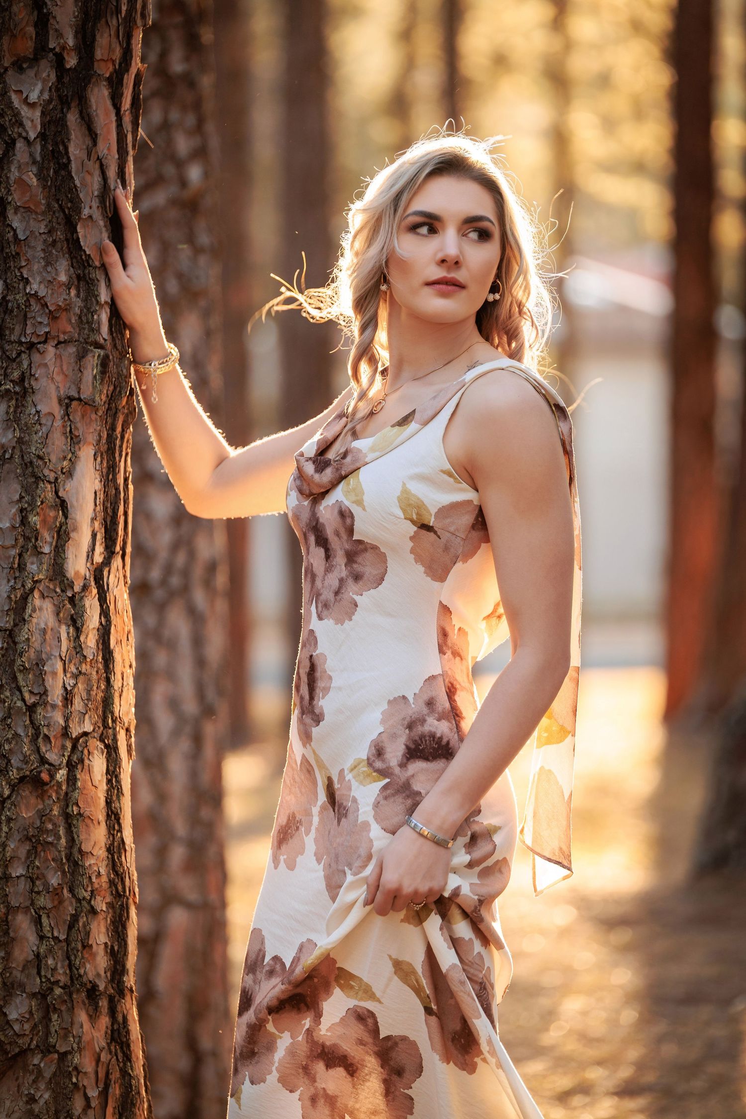 A figure poses against a tree trunk in a sunlit forest wearing a brown and white floral dress at golden hour.