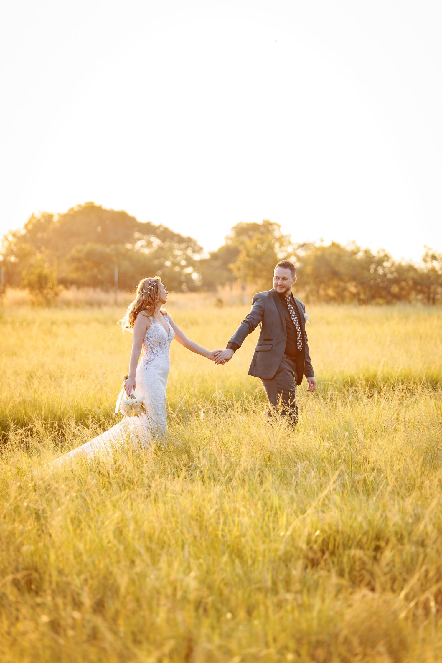 Newlyweds walking through a golden sunlit field during sunset.