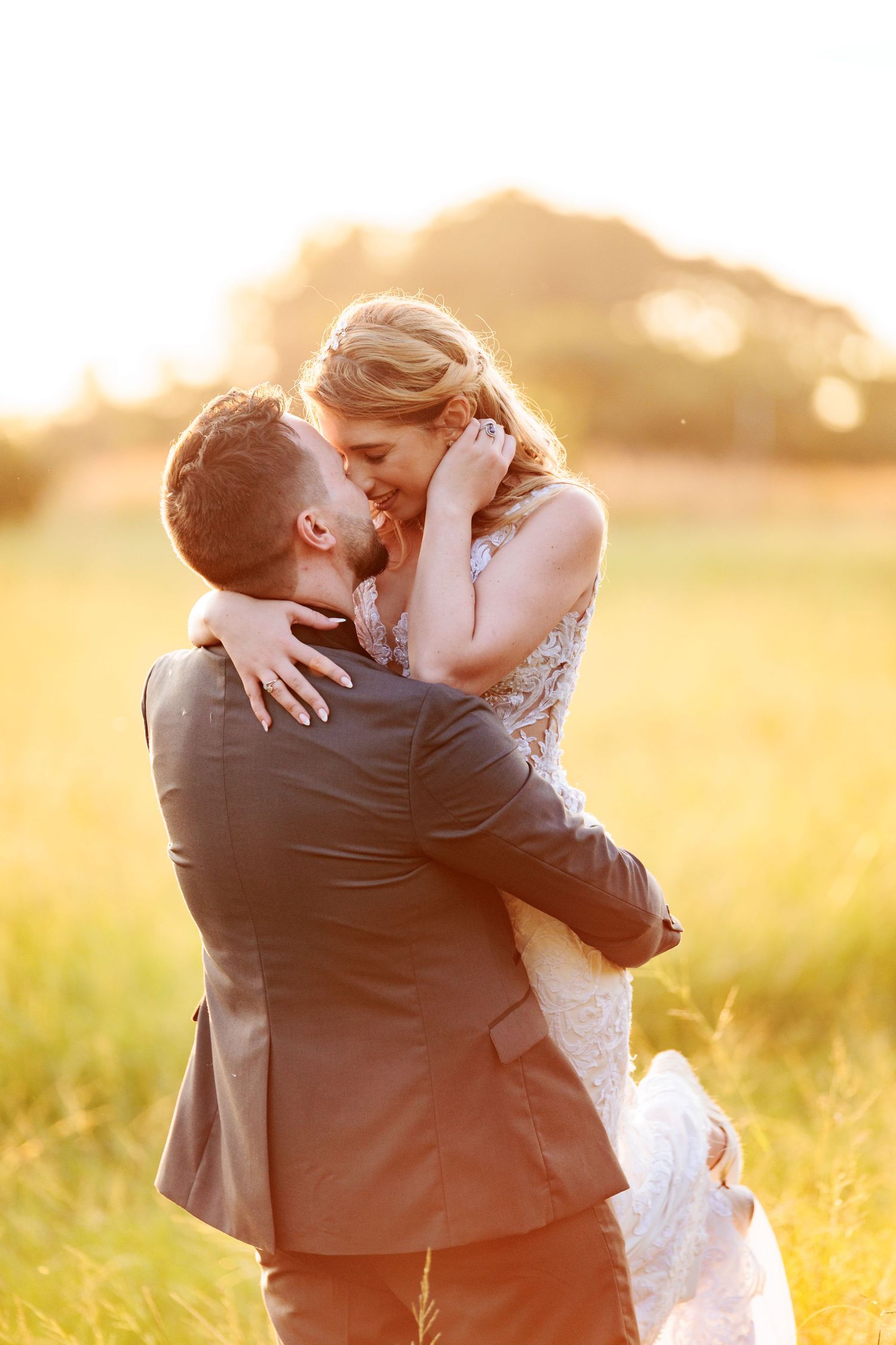 A romantic couple embraces in a sunlit field during golden hour as the sun sets behind them creating a warm glow.