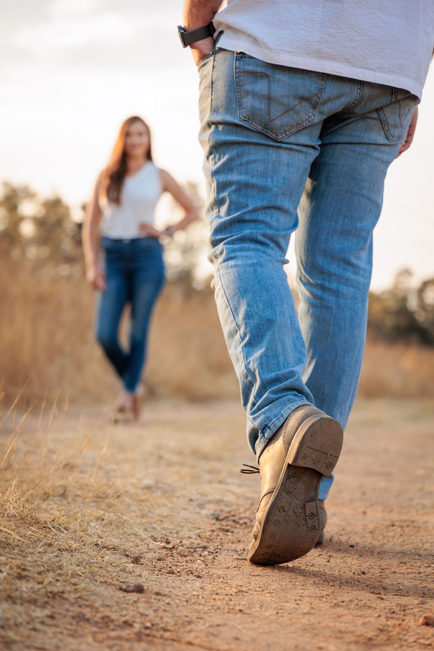 Close-up view of people walking on a dusty dirt path wearing blue jeans and brown boots at sunset.