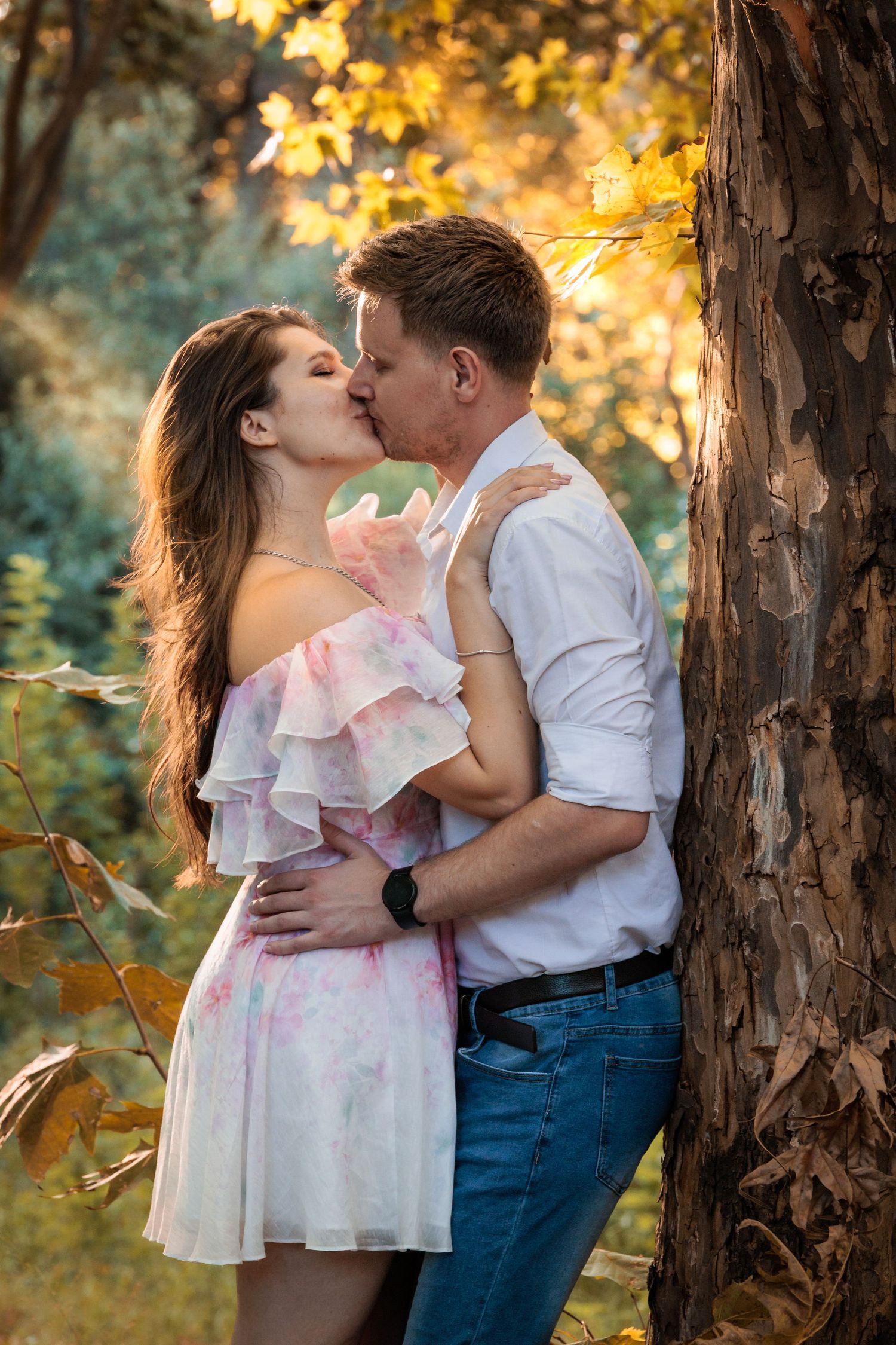 A romantic couple embraces near a tree trunk during golden hour with autumn foliage glowing in the background.