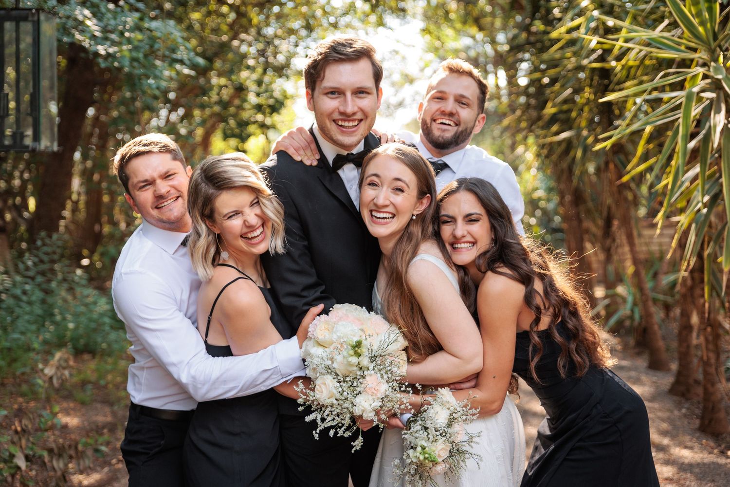 Wedding couple surrounded by smiling friends in formal attire, posing outdoors among lush greenery with bridal bouquet.