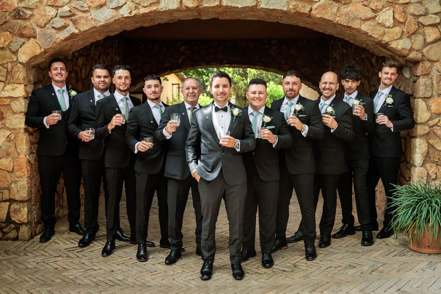 Groom and groomsmen in black tuxedos raising glasses in a toast under a stone arch at a wedding venue.
