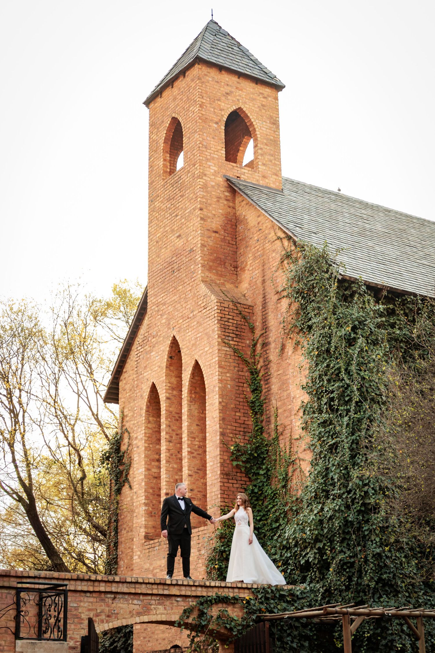 Wedding couple poses together on rustic wooden bridge with historic brick church and ivy-covered walls in background.