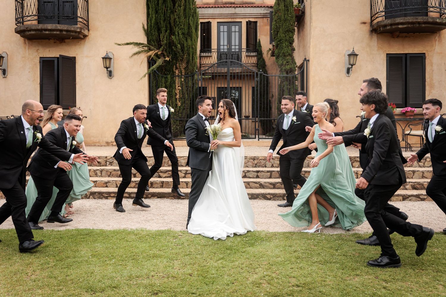 Wedding party members perform a choreographed dance sequence on a lawn in front of a Mediterranean-style venue.