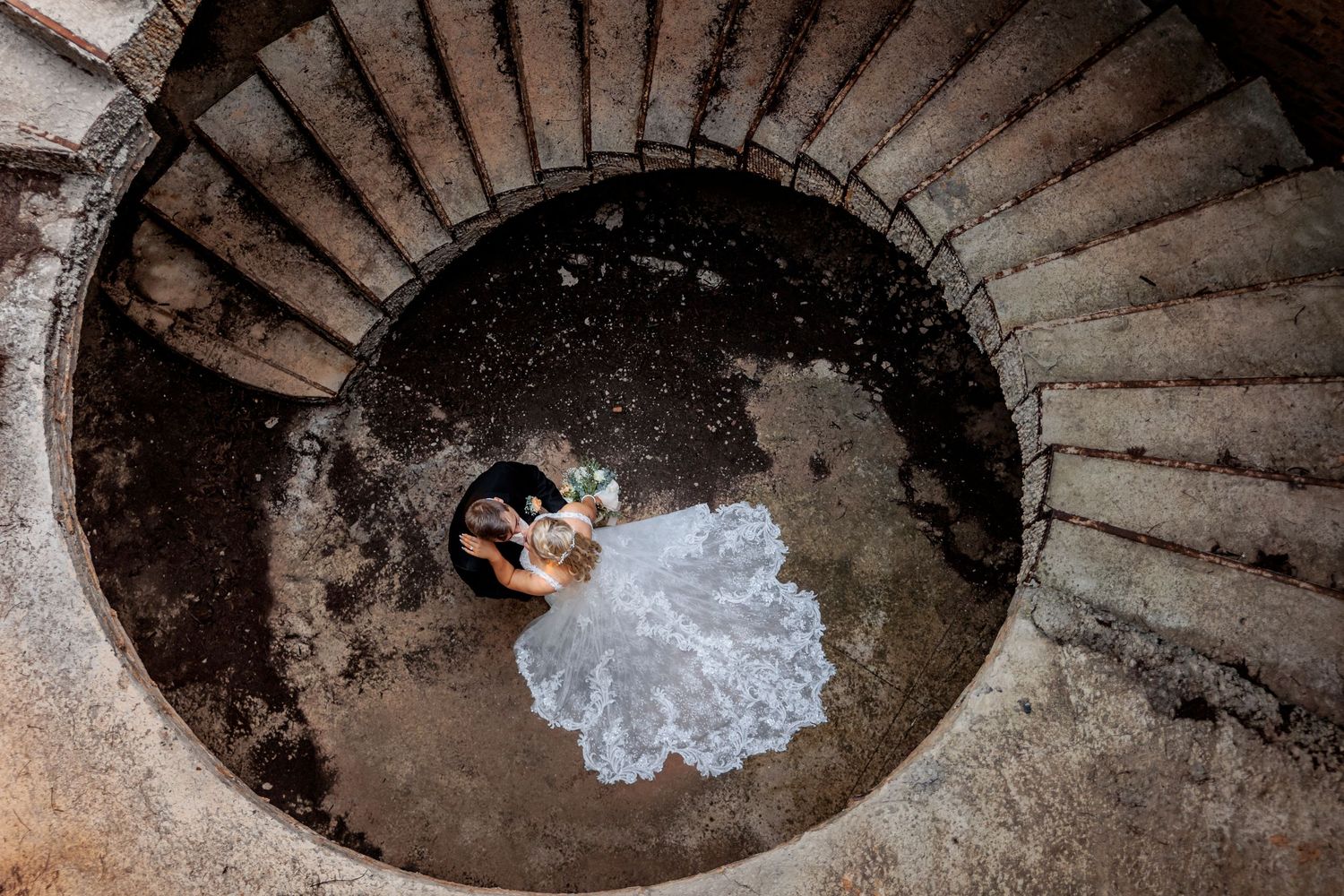 Aerial view of bride and groom embracing at the base of a spiral staircase, with flowing white wedding dress.