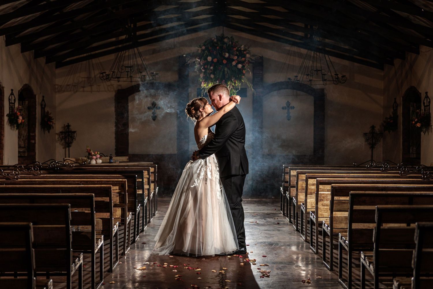 Bride and groom share romantic embrace in rustic chapel aisle, dramatic lighting highlighting their wedding attire.