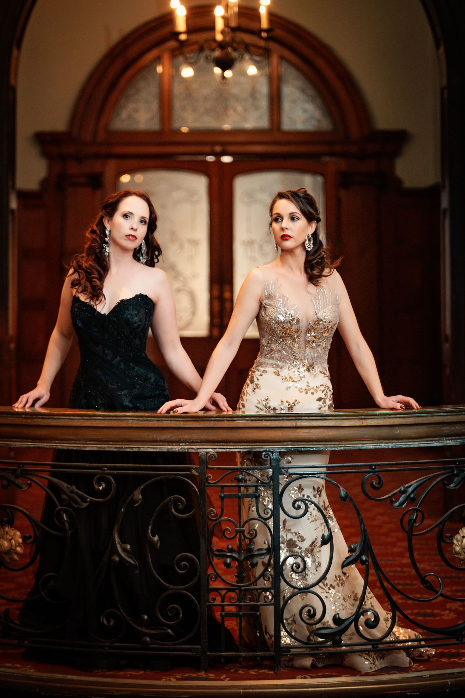 Two women in elegant evening gowns pose together on an ornate balcony with wrought iron railings in a vintage setting.