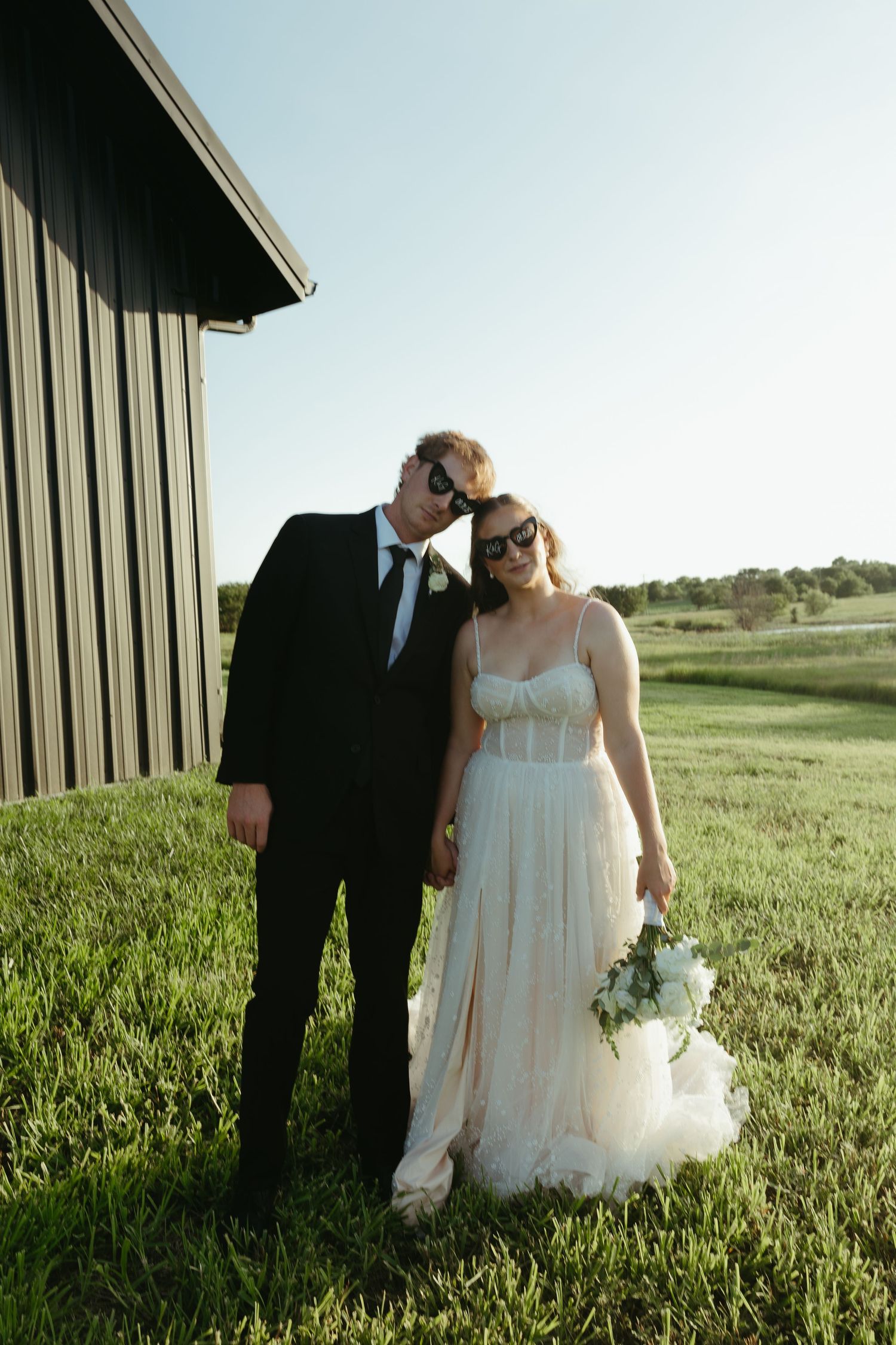 Wedding couple embraces in a green field next to a modern barn building.