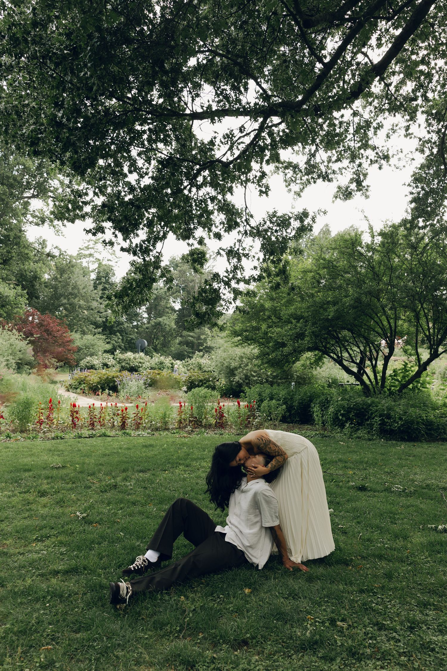Couple sitting together on green grass in a lush garden park, sharing a tender romantic moment under a large tree.