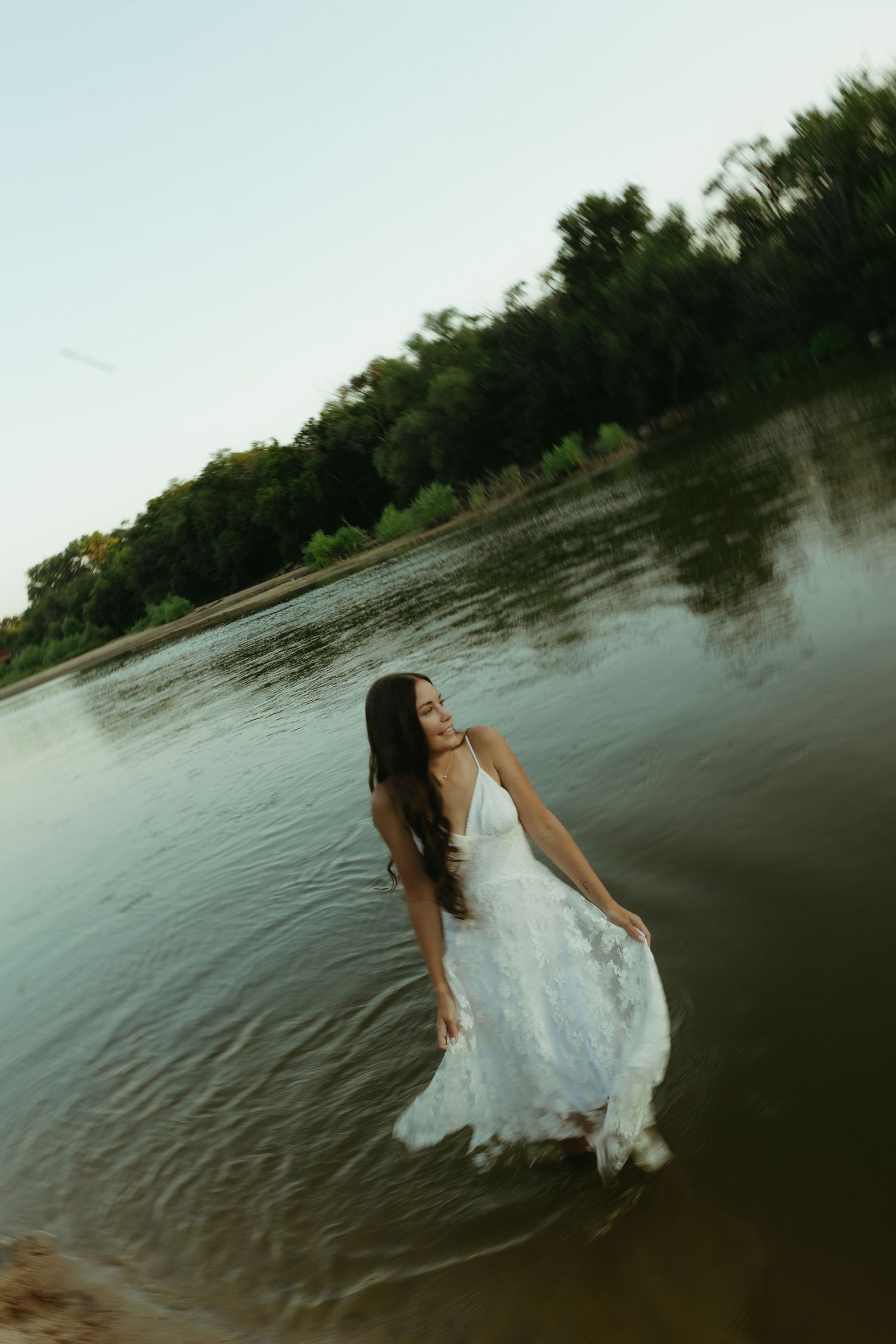 A woman in a flowing white dress stands gracefully in a calm river, surrounded by lush green trees in soft evening light.