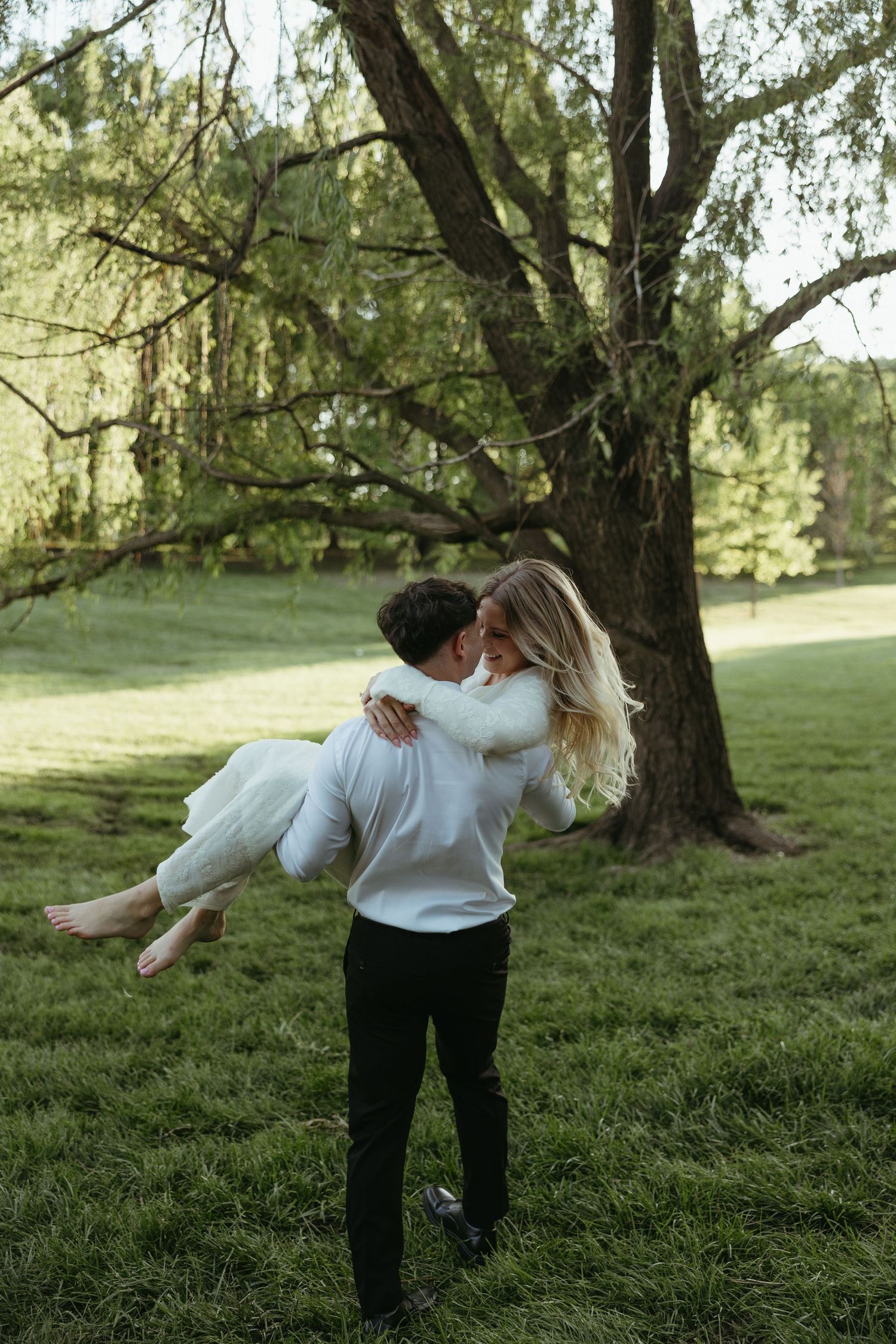 Groom carries bride in white dress outdoors near large tree on lush green grass in romantic wedding photo.