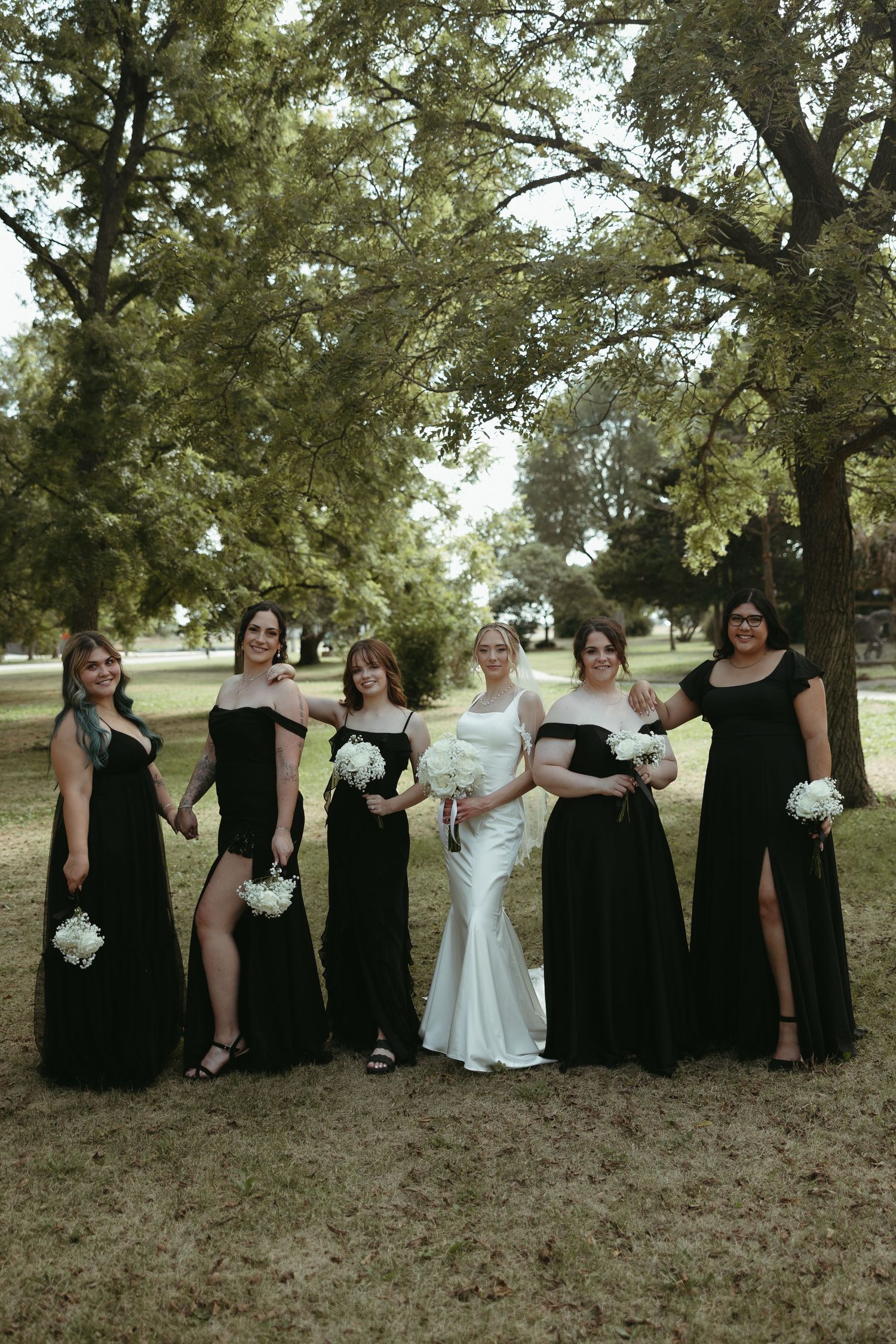 Wedding party in black dresses poses together under trees for group photo.