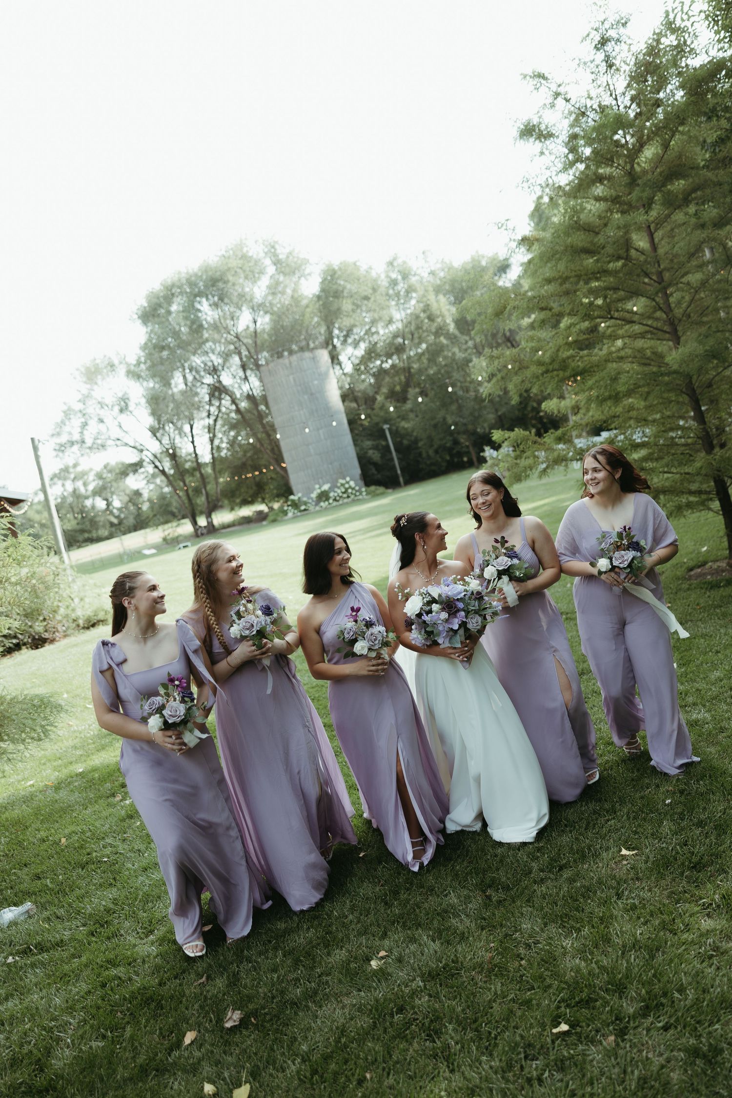 Bridesmaids in lavender dresses pose together on grassy lawn during outdoor wedding.