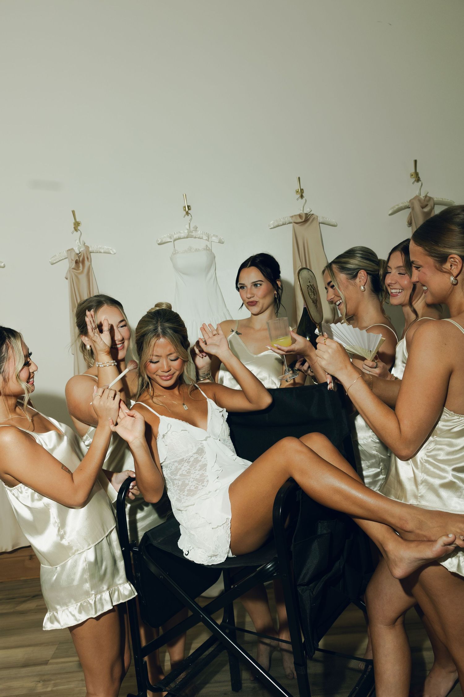 Bridesmaids in satin slip dresses celebrate with the bride, laughing and toasting with cocktails during bridal party preparations.