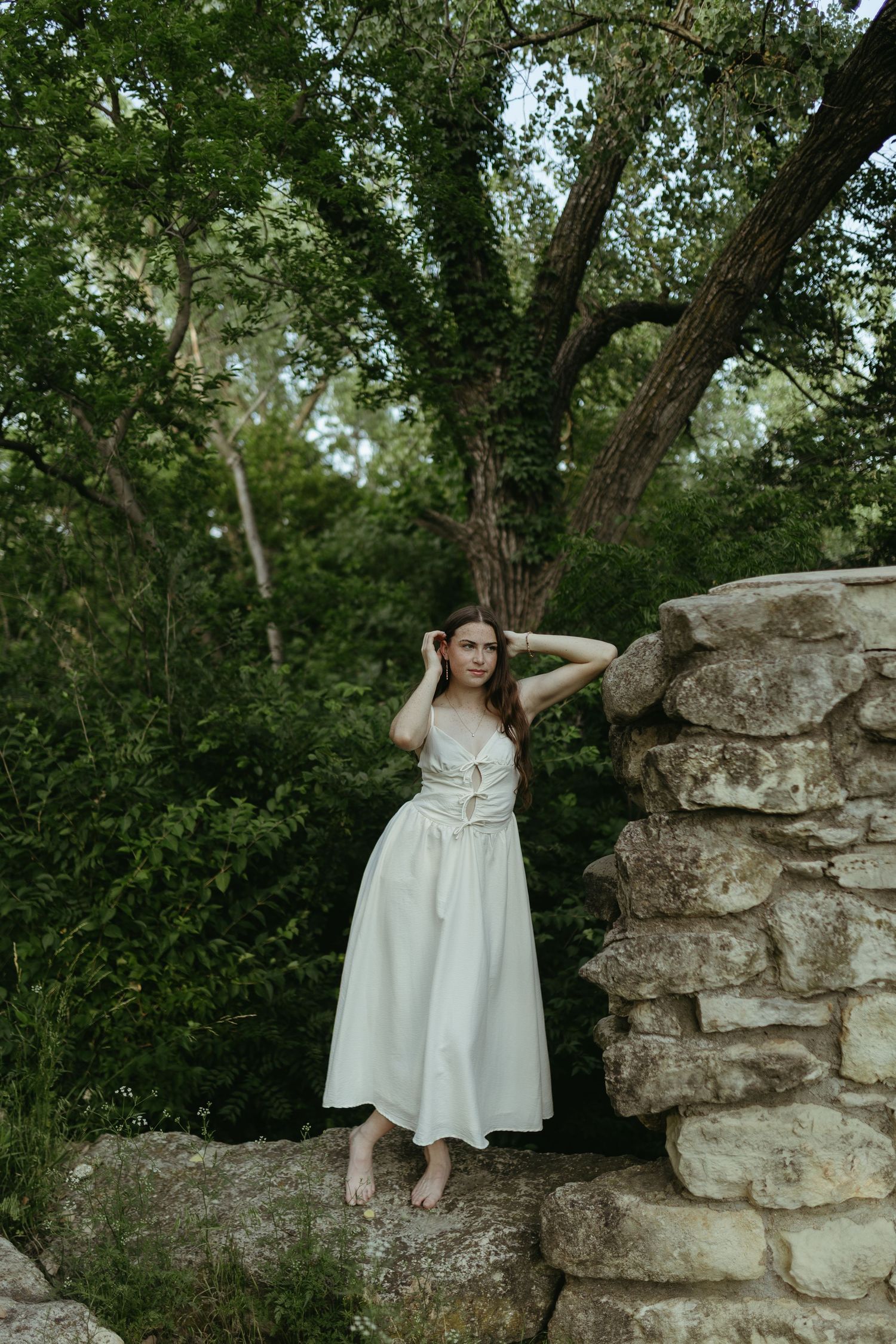 Woman in white dress poses by stone wall surrounded by lush green trees in an outdoor natural setting.