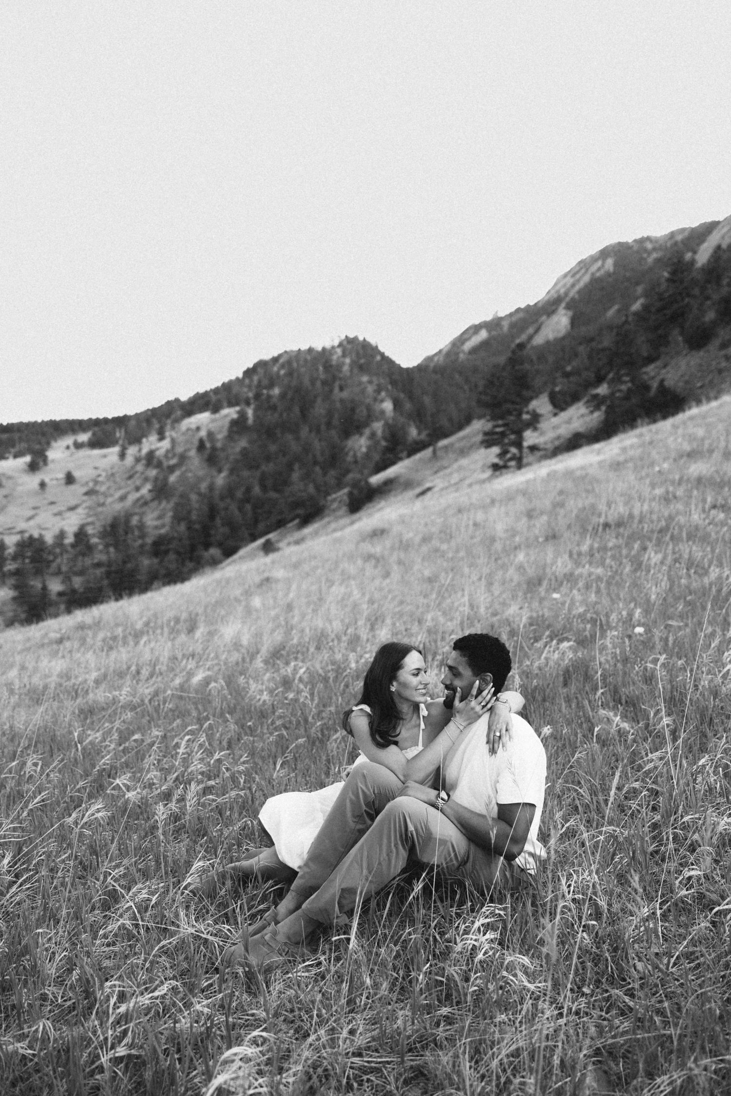 Black and white photo of a romantic couple sitting in a grassy mountain meadow, gazing at each other tenderly.