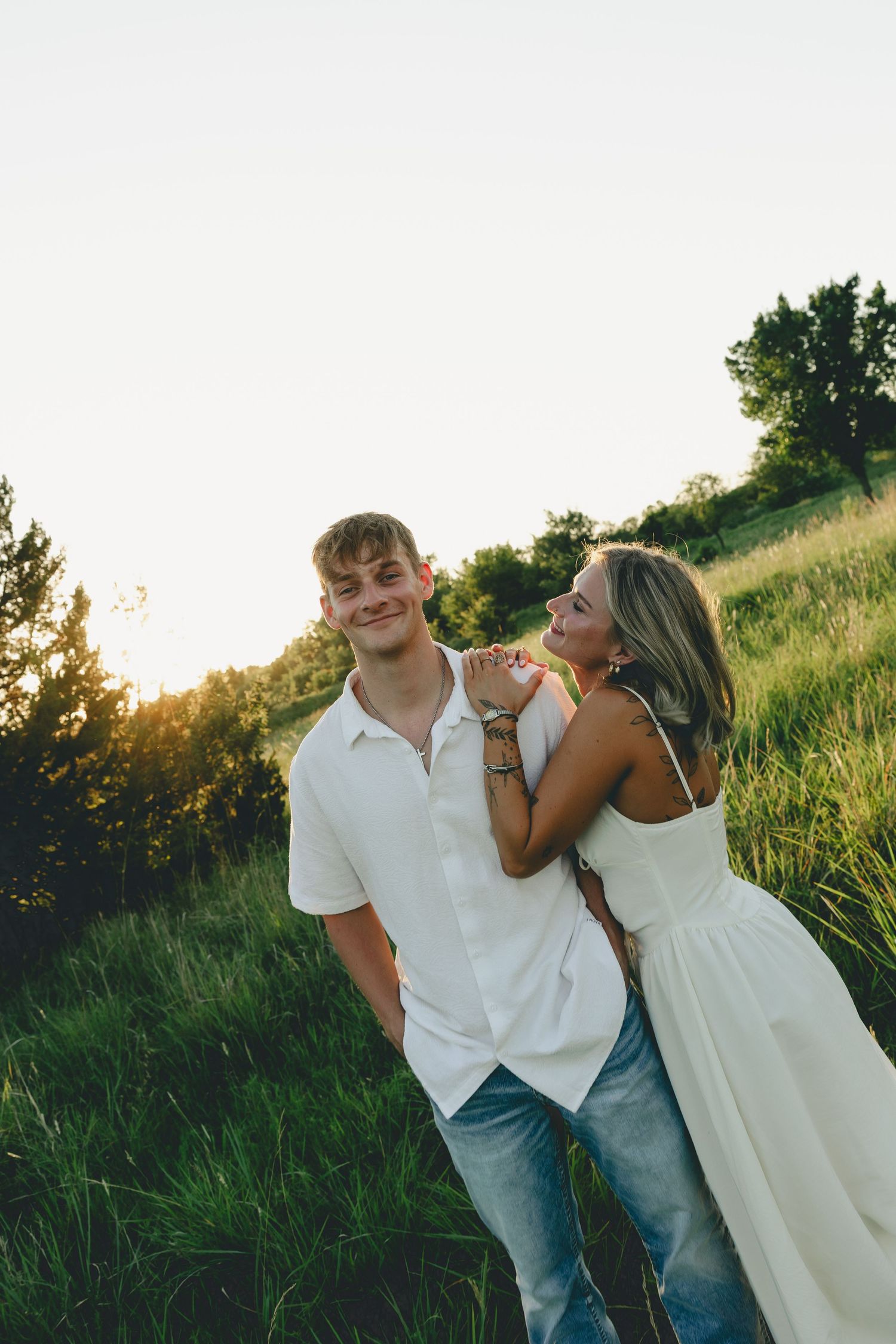 A young couple in white outfits poses in a sunlit green field, the woman leaning affectionately on the mans shoulder.
