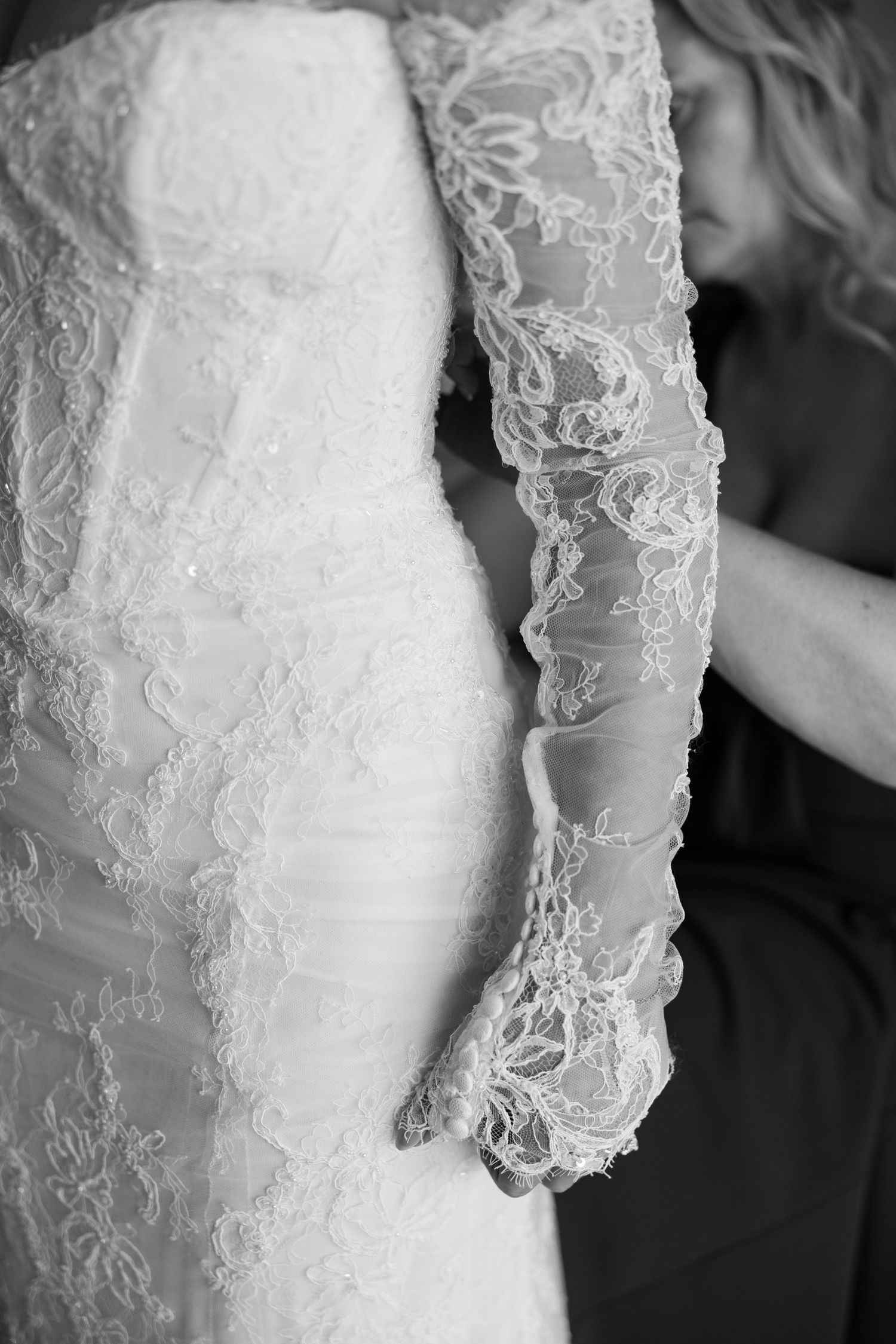 Black and white close-up of a bride in a lace wedding gown with long lace sleeves being buttoned up.
