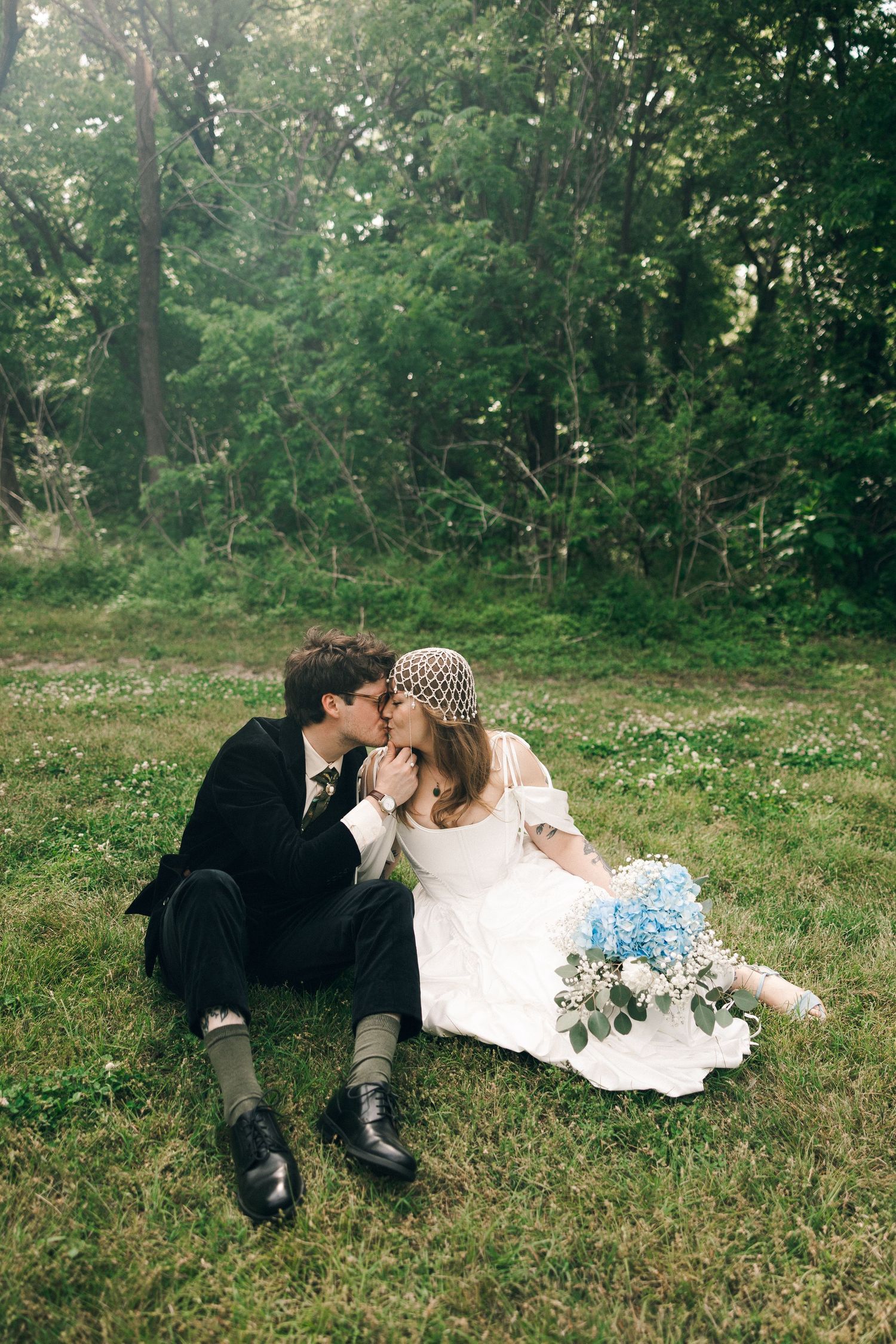Bride and groom kissing while sitting on green grass outdoors, bride holding blue hydrangea bouquet, surrounded by lush trees.