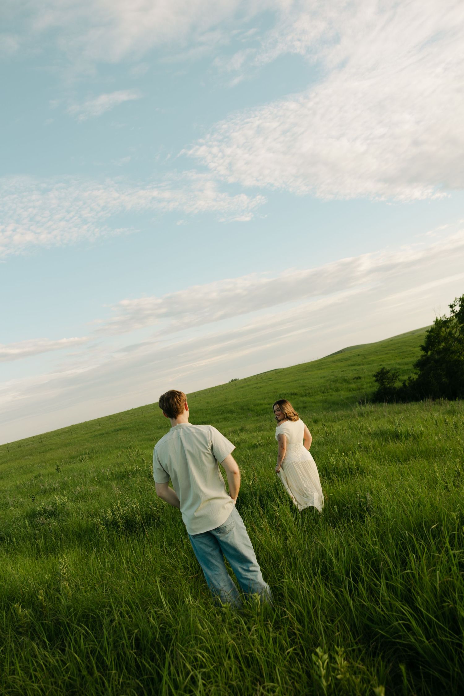 A couple walks through a lush green meadow under a partly cloudy sky, the woman in a white dress leading the way.