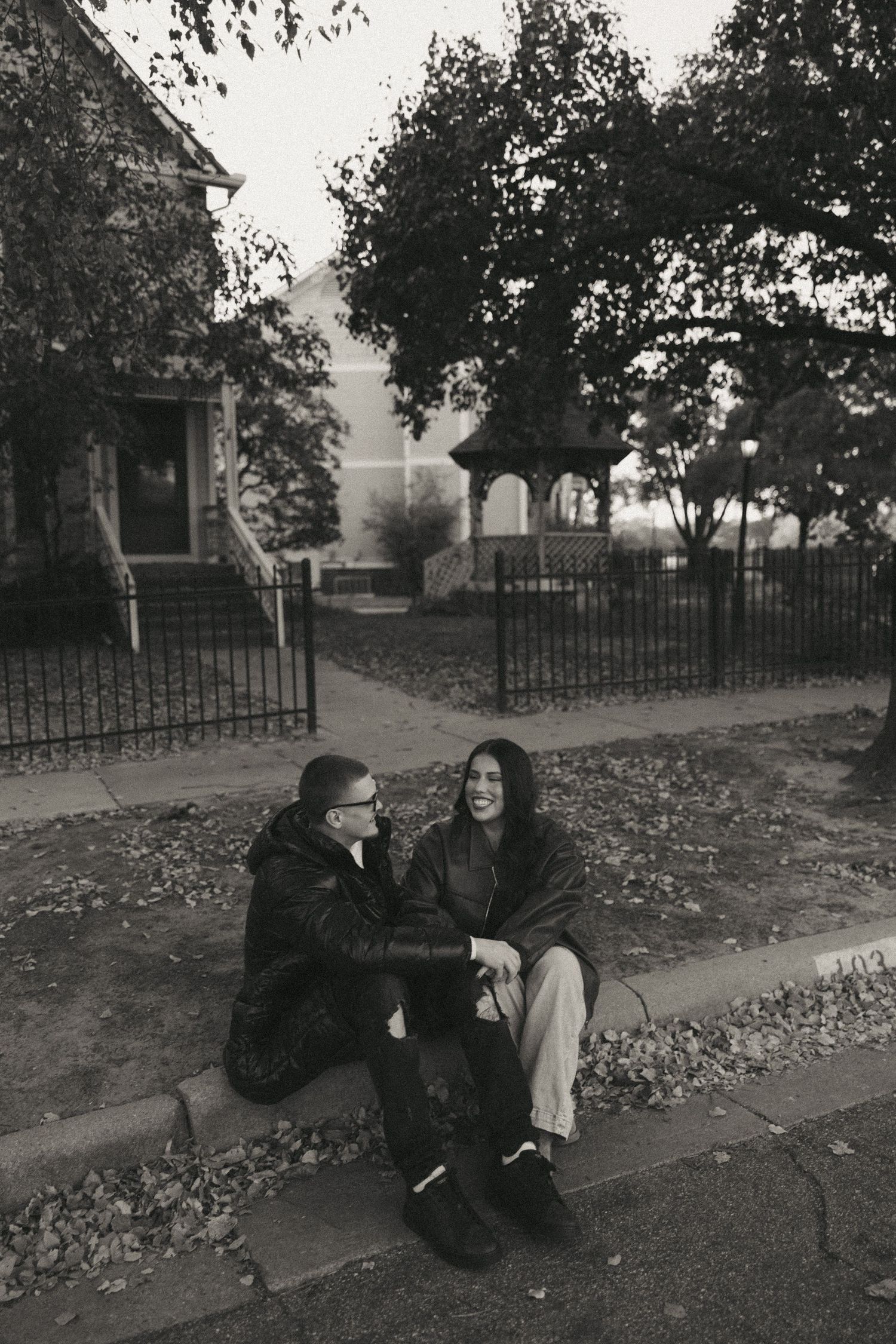 Black and white photos of a couple sitting and laughing together on grass near an iron fence.