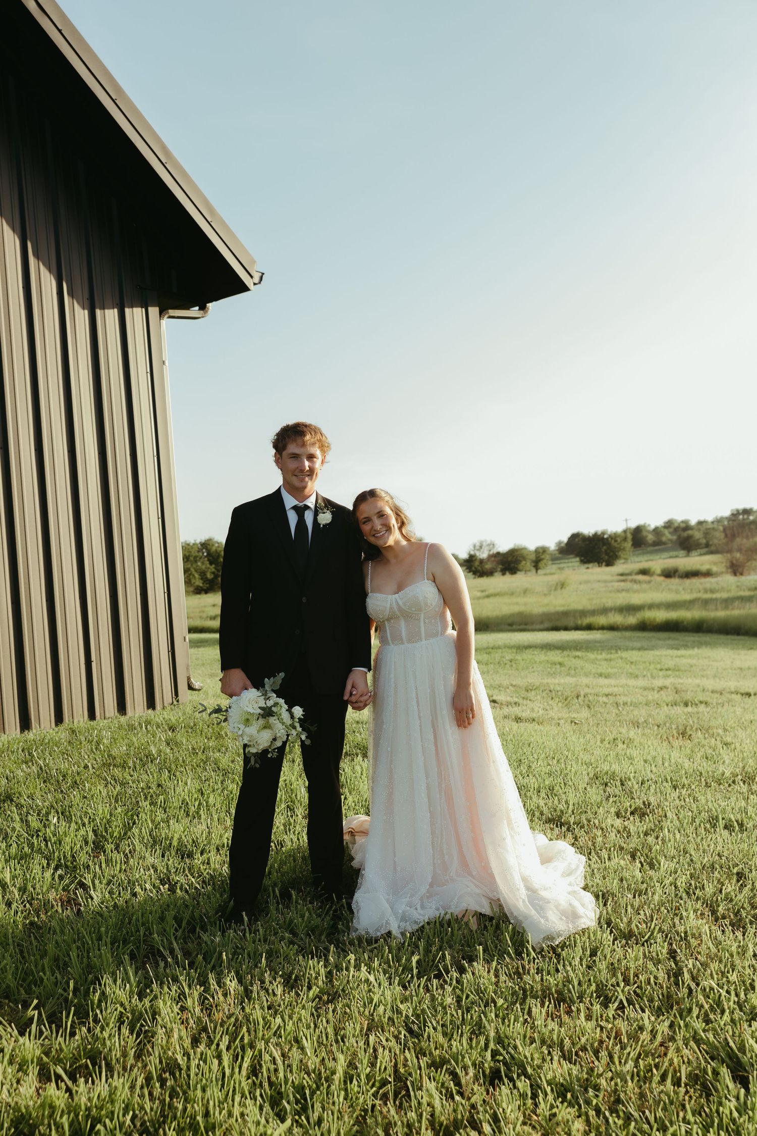 Newlyweds pose together in a sunlit meadow beside a rustic barn structure.