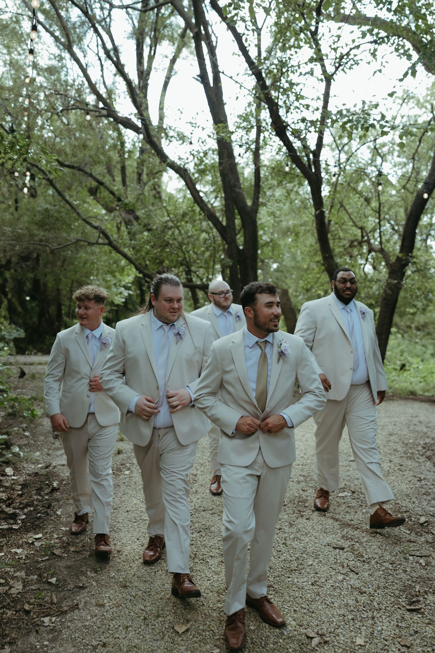 Groomsmen in light colored suits walk together on outdoor path.
