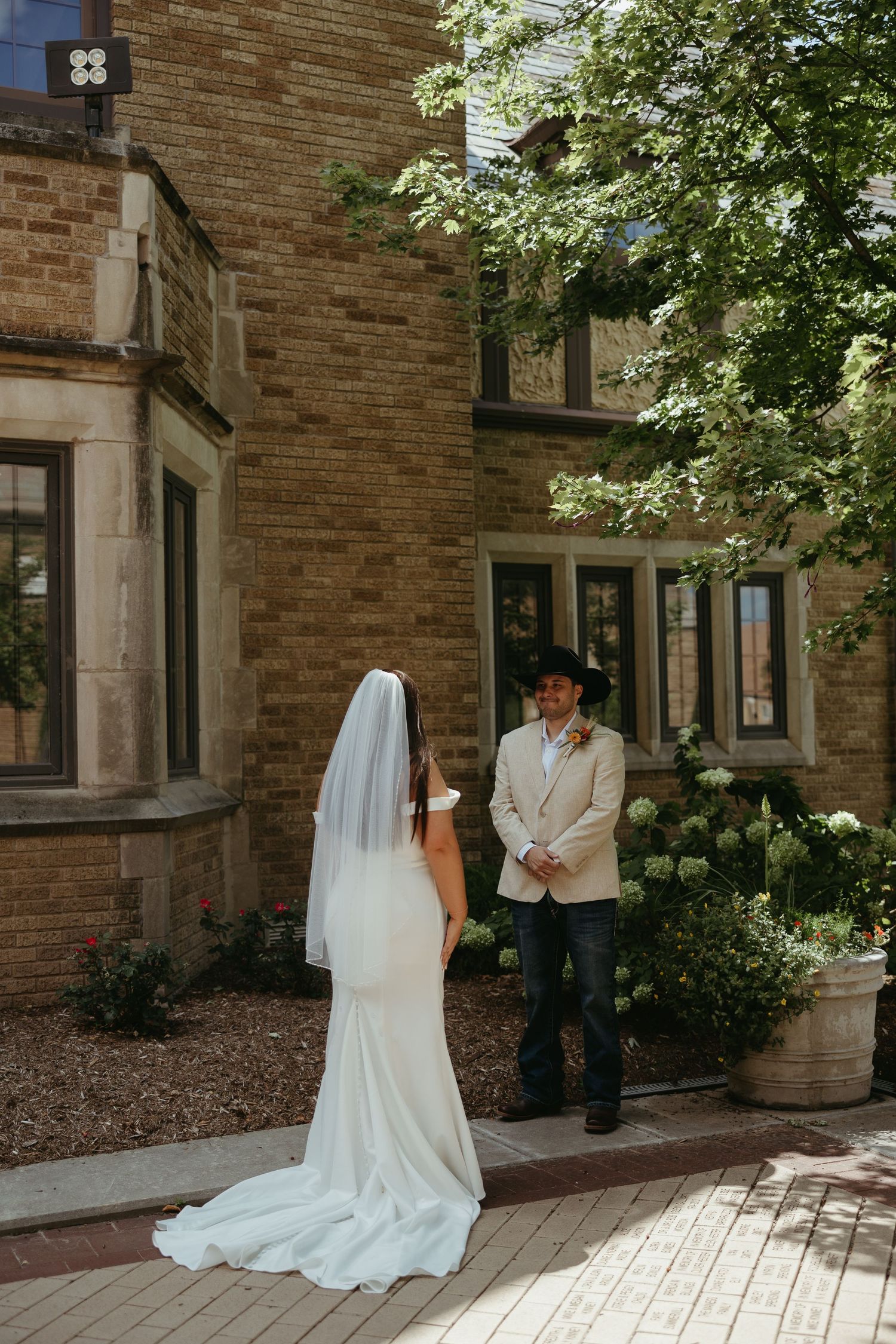 A wedding first look moment captured in a brick courtyard with sunlight streaming through trees.