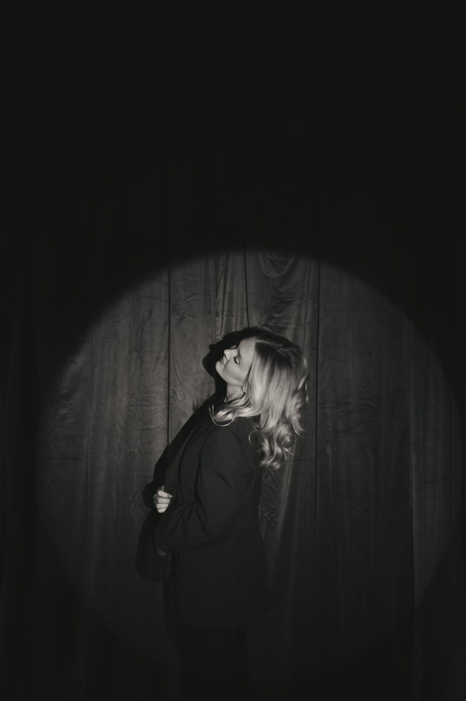 Black and white portrait of a blonde woman in dark clothing, illuminated by a circular spotlight against a wooden backdrop.