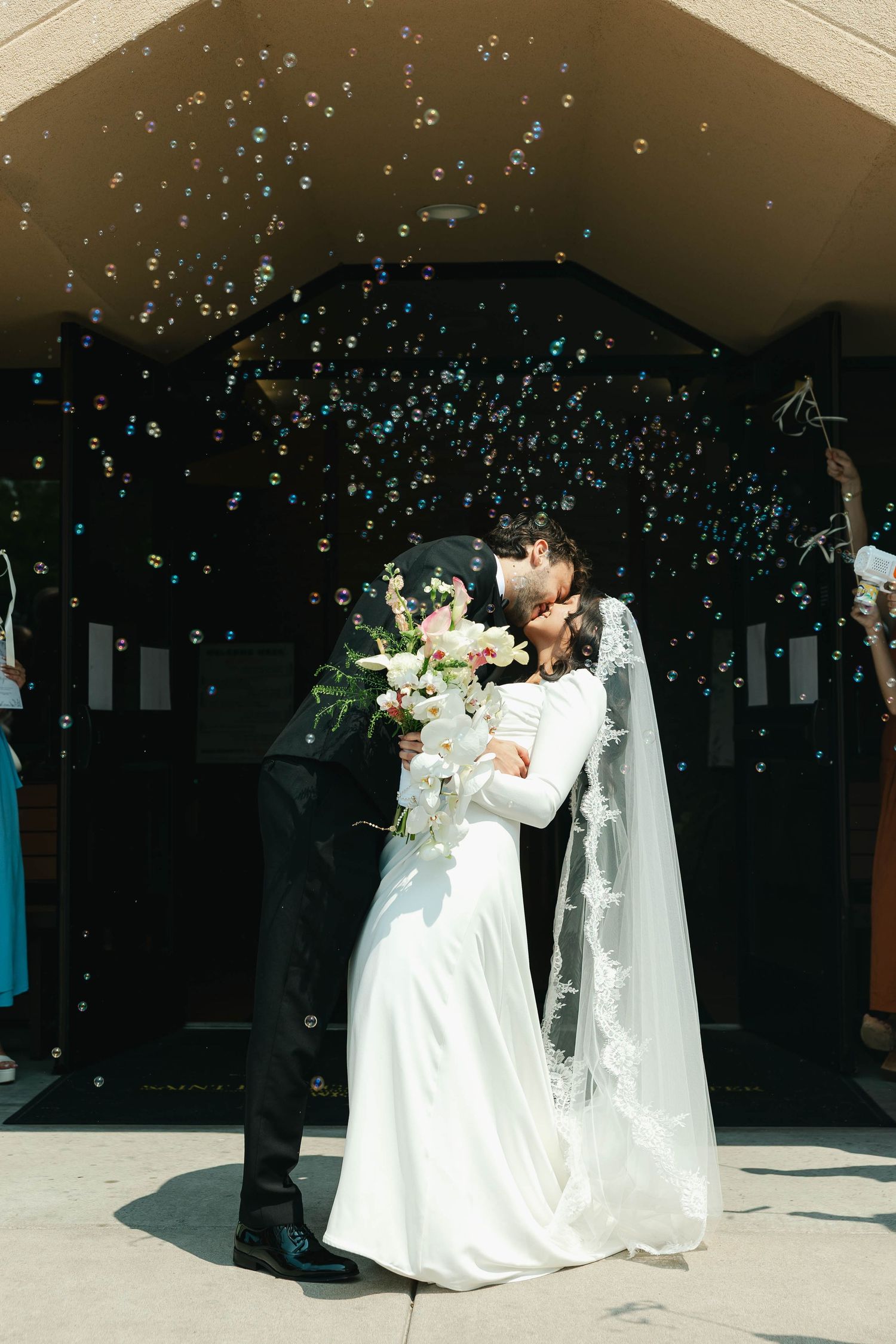 Newlywed couple sharing a kiss outside church entrance surrounded by floating bubbles, bride in white gown holding bouquet.
