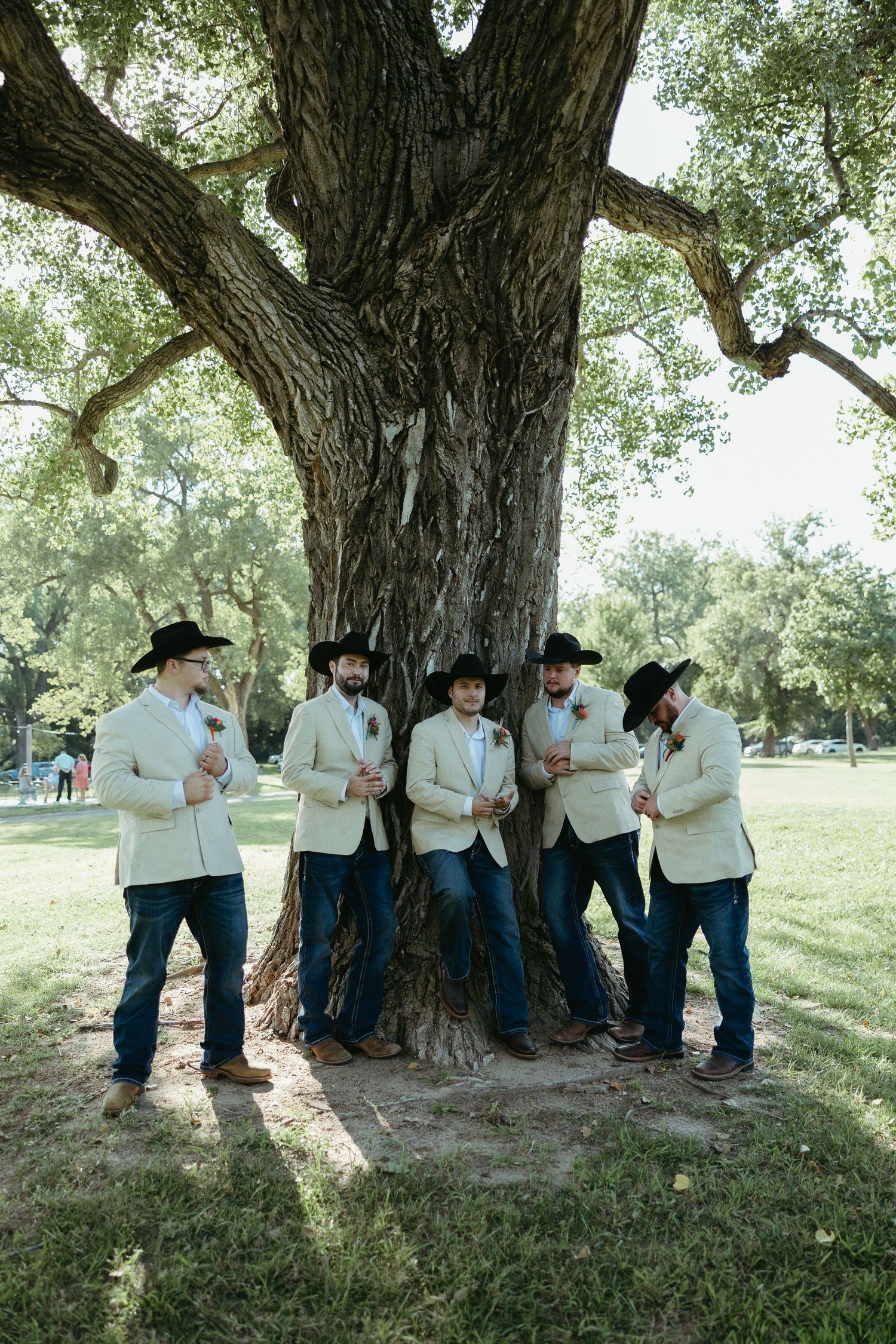 Groomsmen in white shirts and cowboy hats pose casually under a large tree.