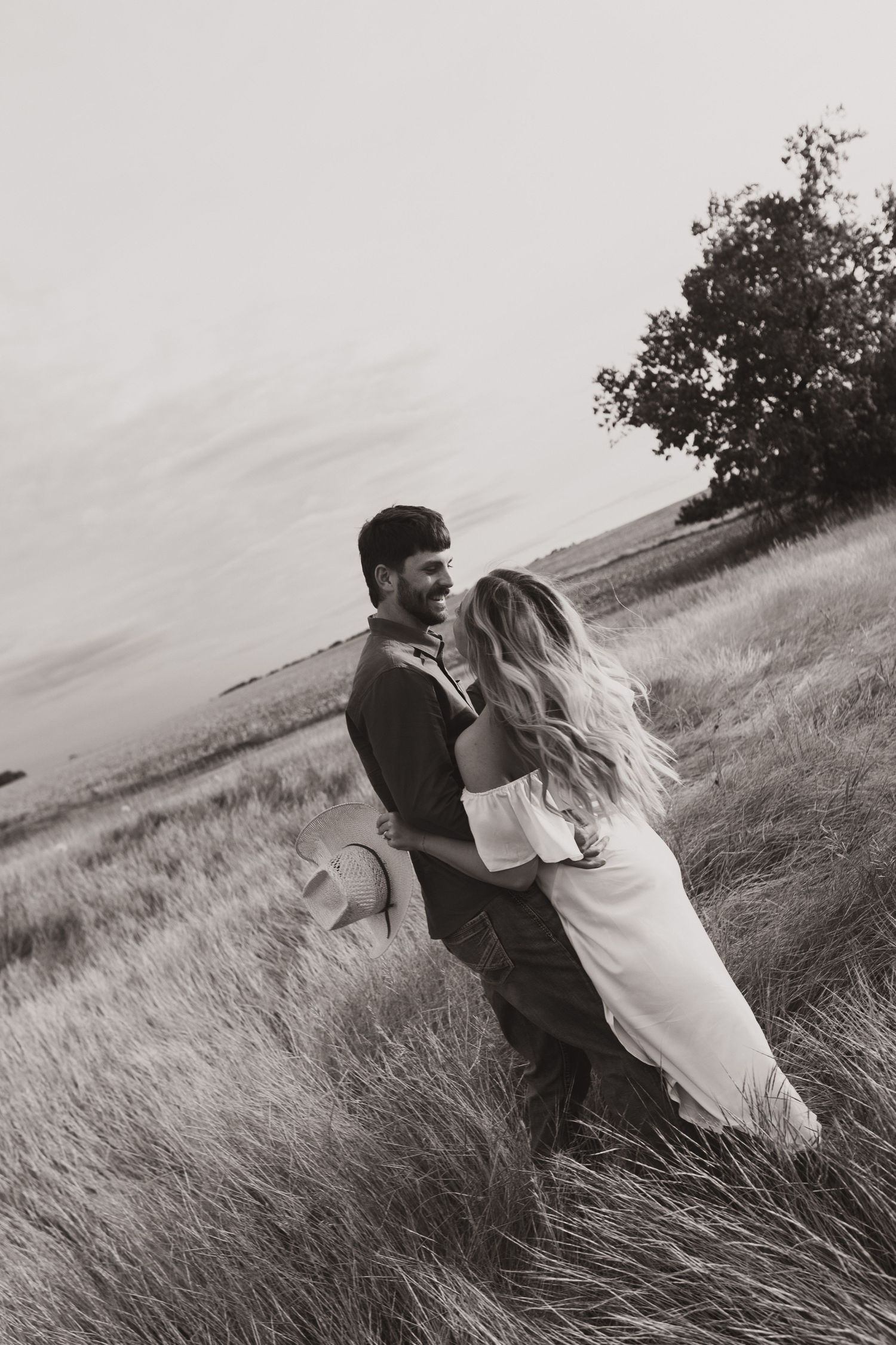 Black and white photo of a couple embracing romantically in a golden wheat field with a tree in the background.