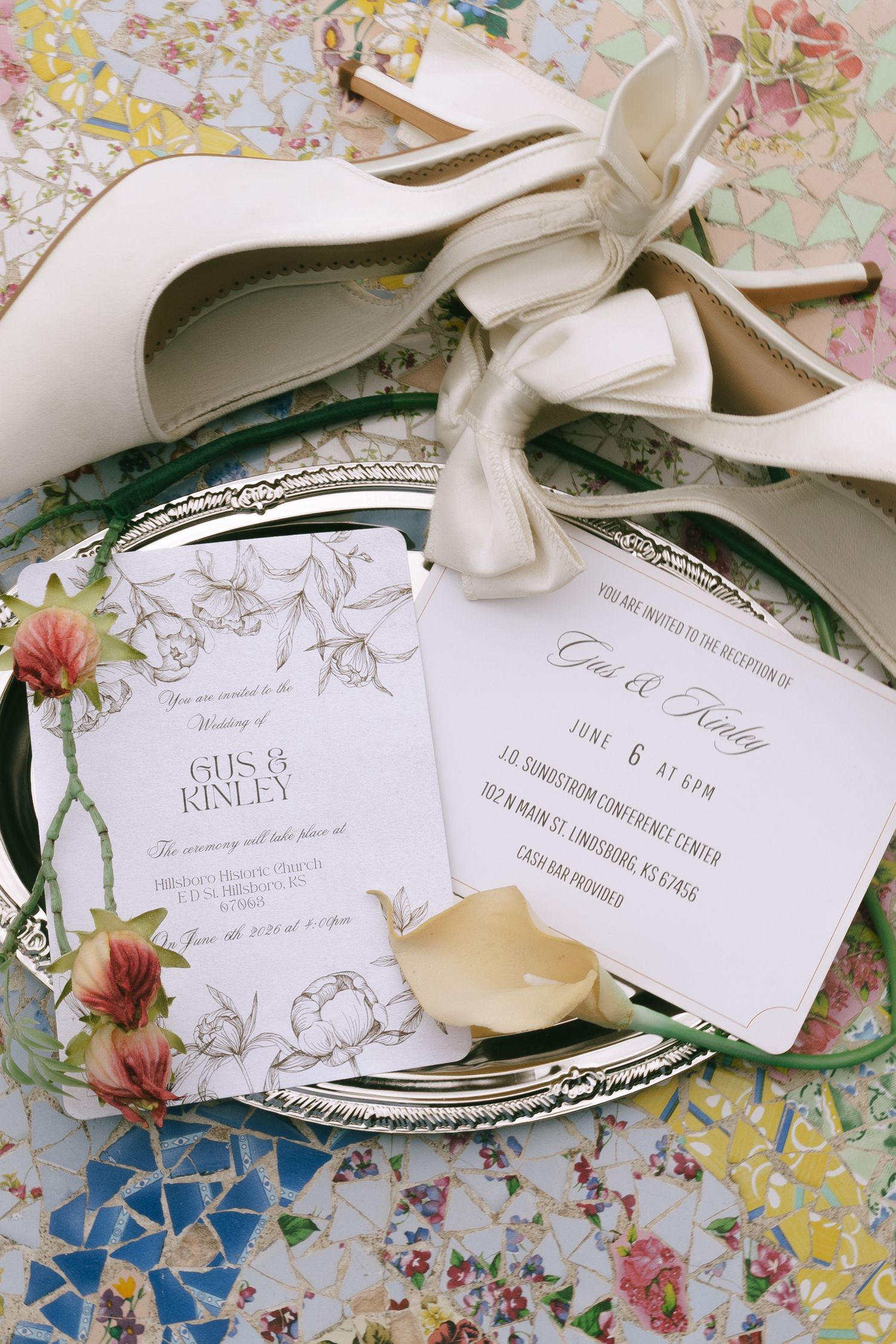 Elegant wedding invitation suite for Gus and Kinley displayed on silver tray with white heels on colorful mosaic surface.
