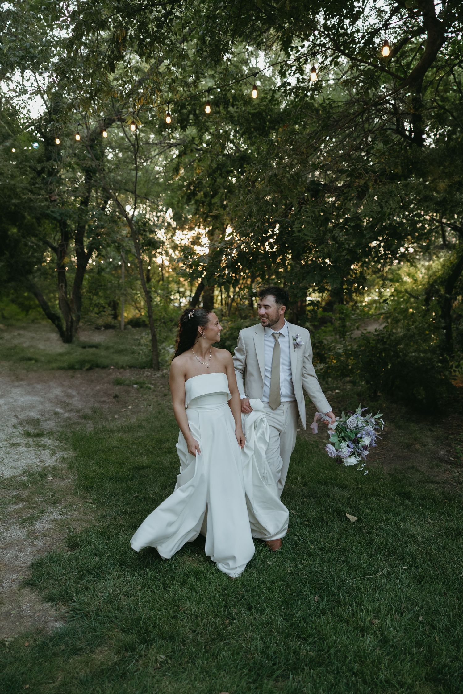 Couple in wedding attire walks through green garden setting at sunset.