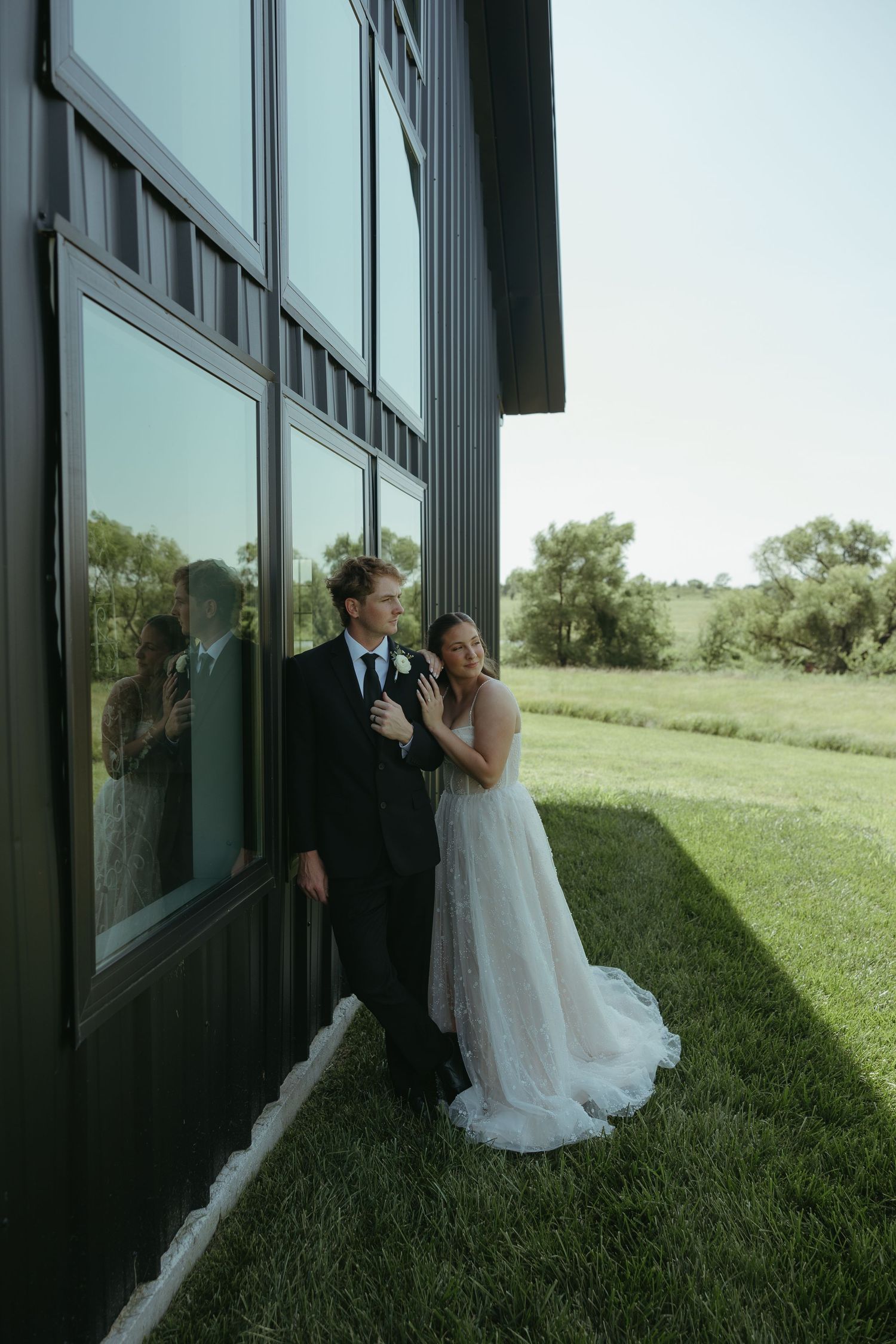 Newlyweds pose against a modern glass building reflecting the green landscape.