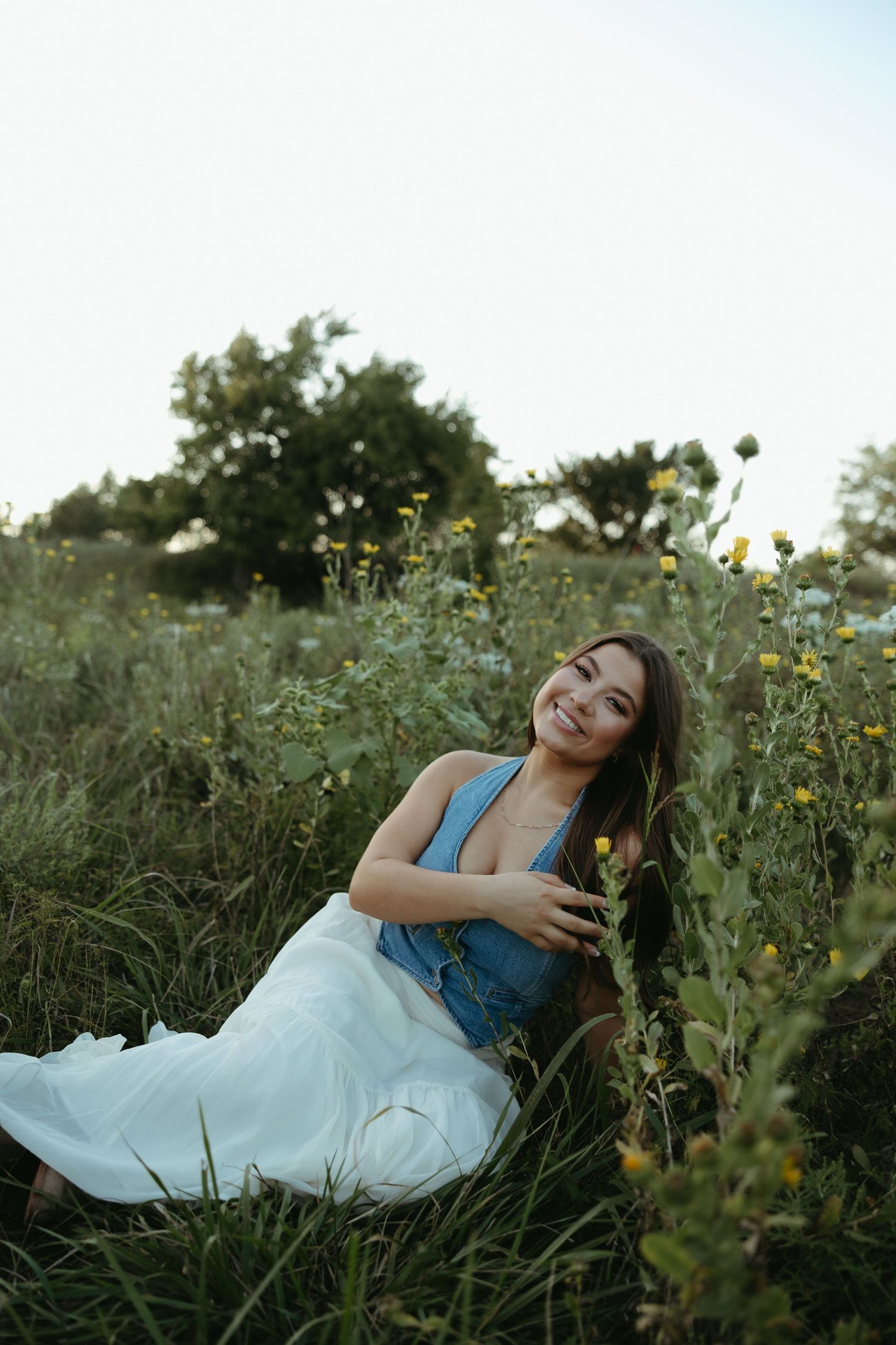 Smiling young woman in blue top and white skirt sitting among wildflowers in a lush green field.