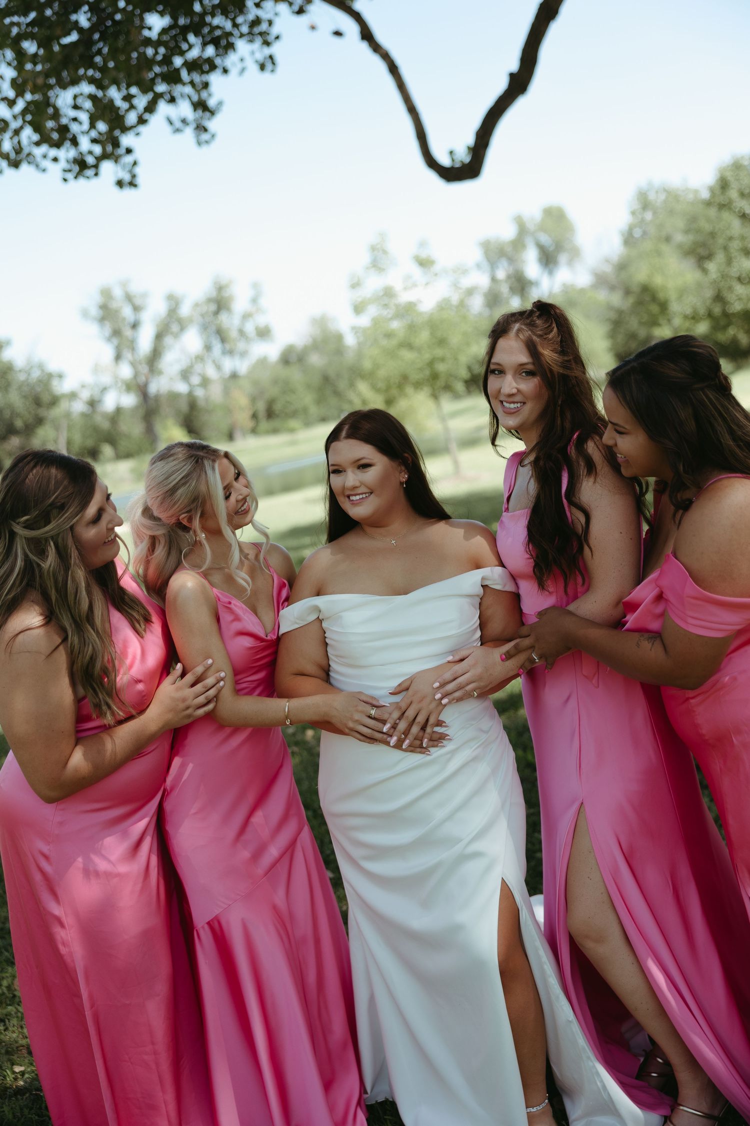 Bridesmaids in pink dresses gather around the bride for a celebratory group hug outdoors.