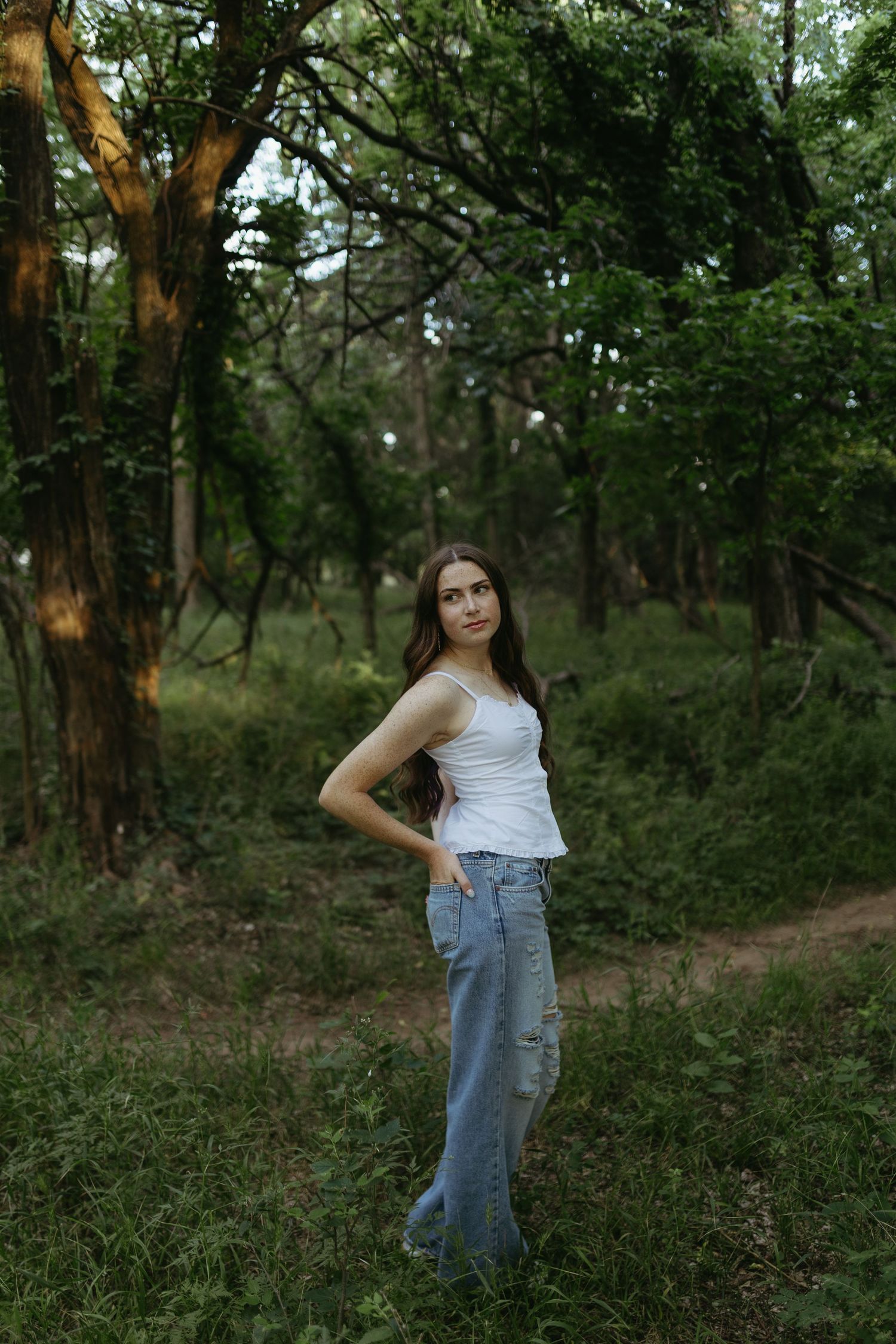 Young woman in white tank top and ripped jeans poses confidently in a lush green forest setting.