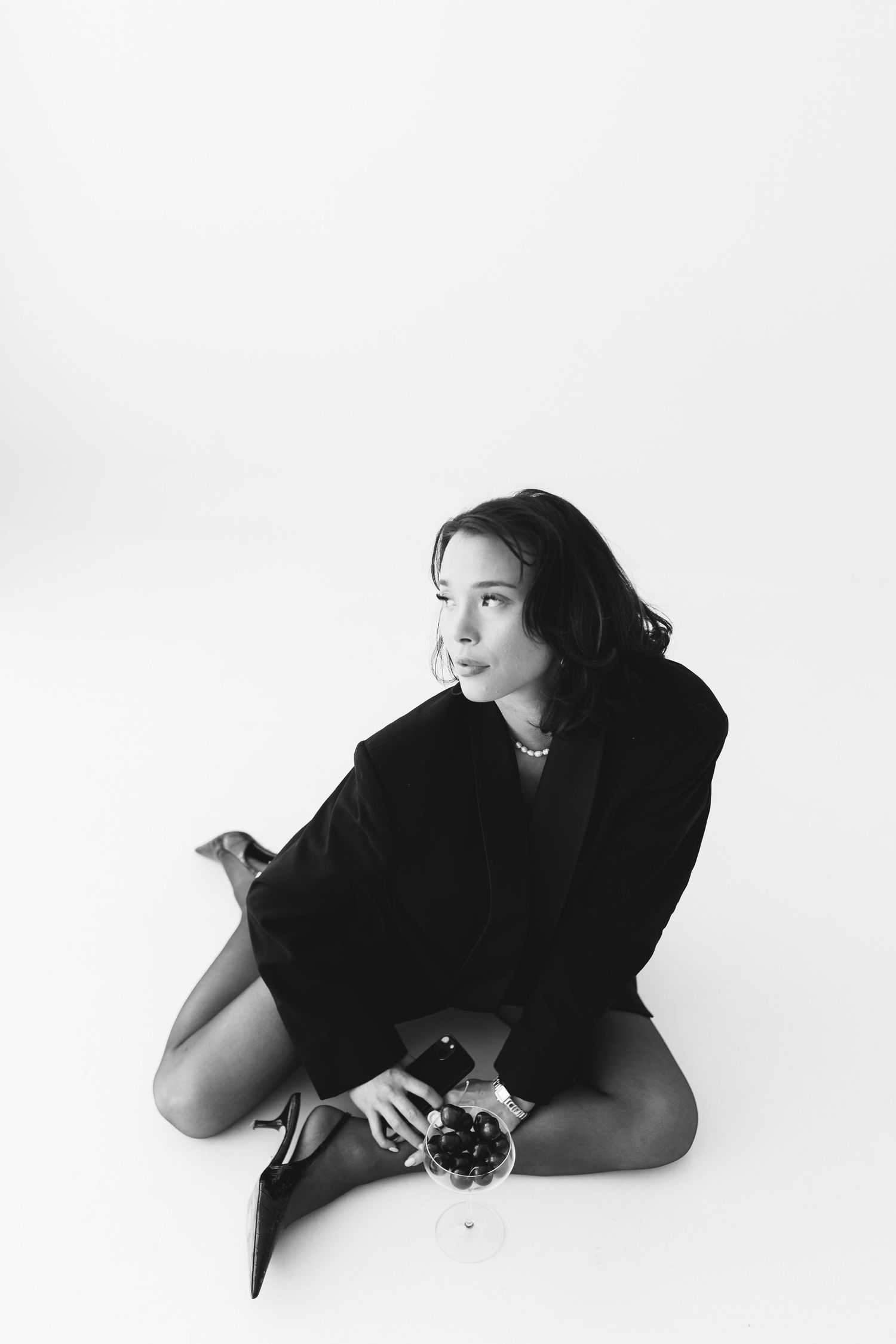 Black and white photo of a young woman in a blazer sitting on the floor holding a wine glass with grapes.