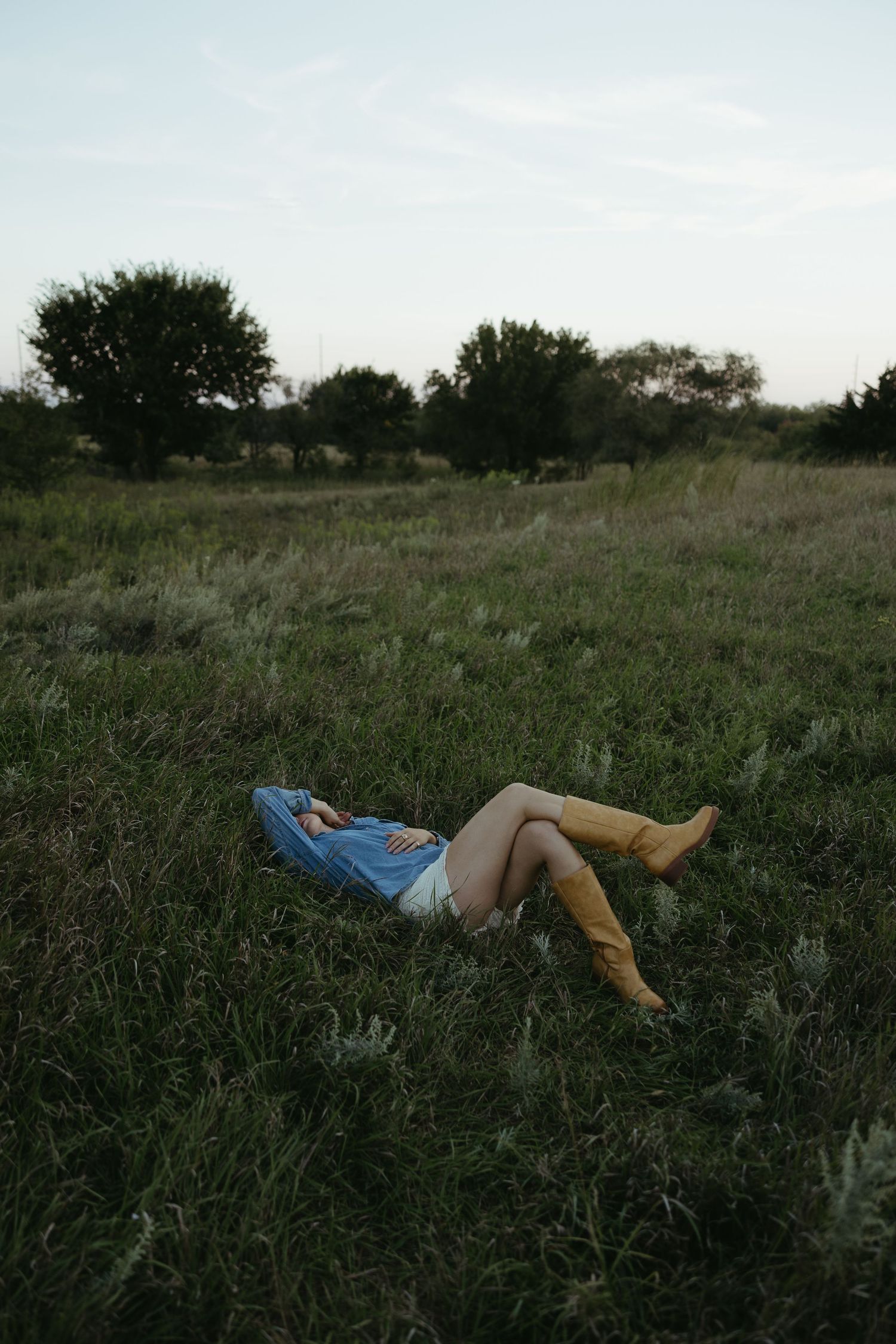 A relaxed figure lies in tall grass during golden hour wearing cowboy boots and a blue dress.