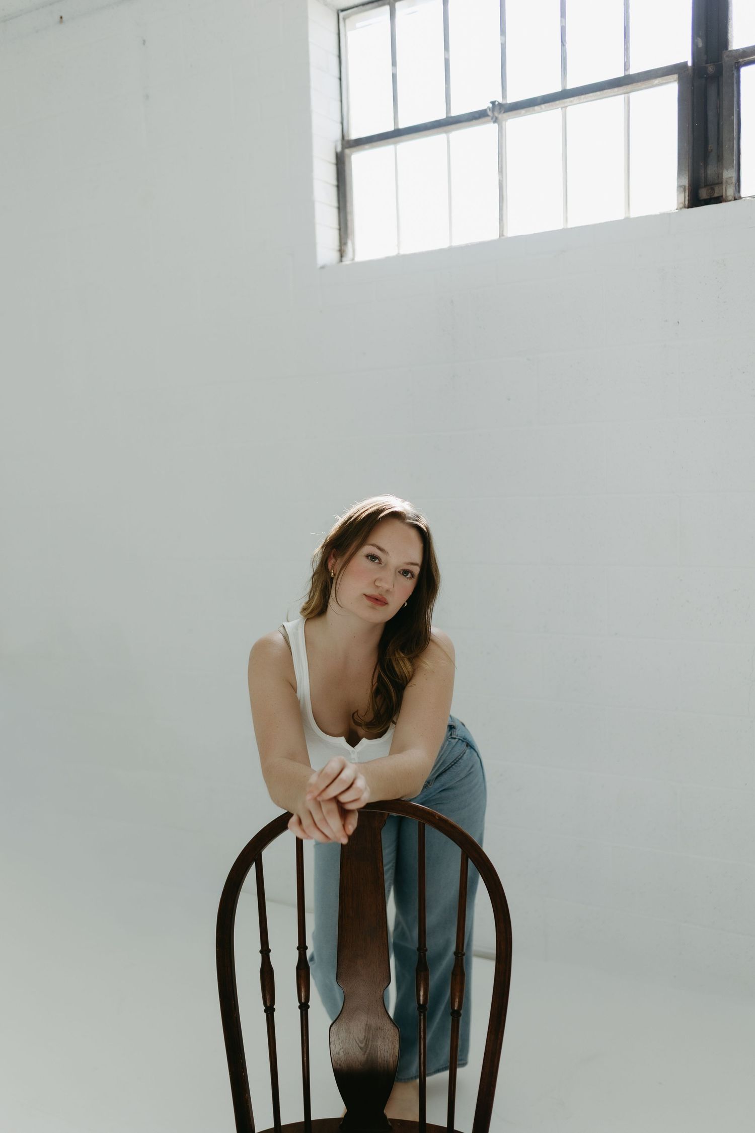 Young woman in white top and blue skirt sits backwards on wooden chair in bright minimalist studio.