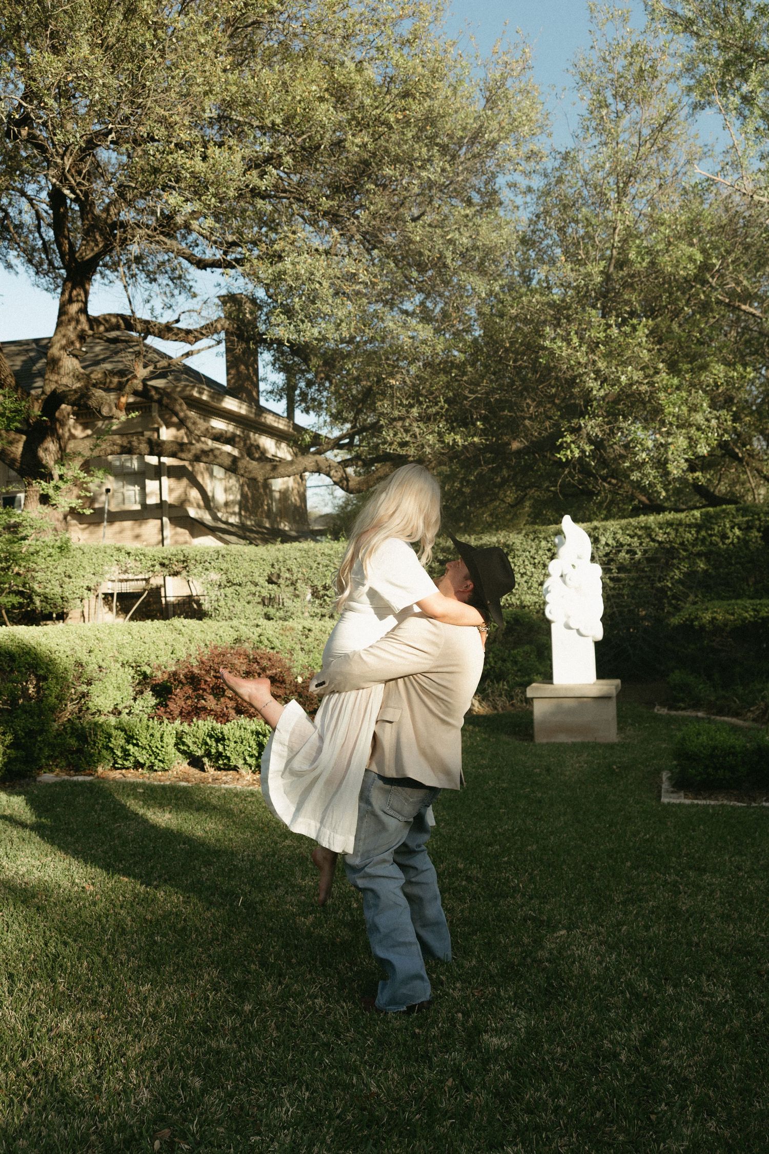A man lifts a woman in a romantic garden setting with a white statue and lush greenery in the background.